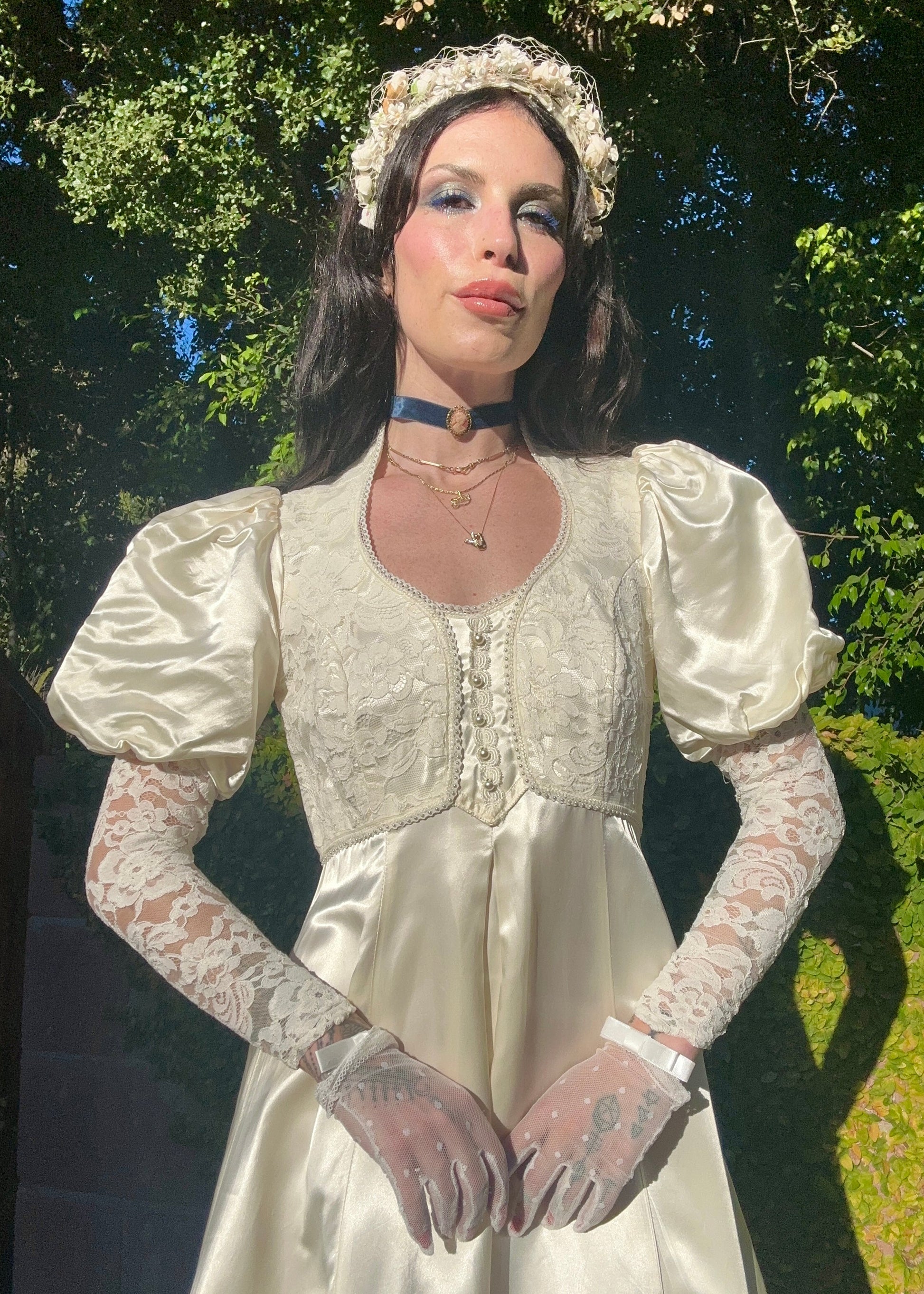 Woman in a white vintage dress with lace sleeves and gloves standing outdoors.