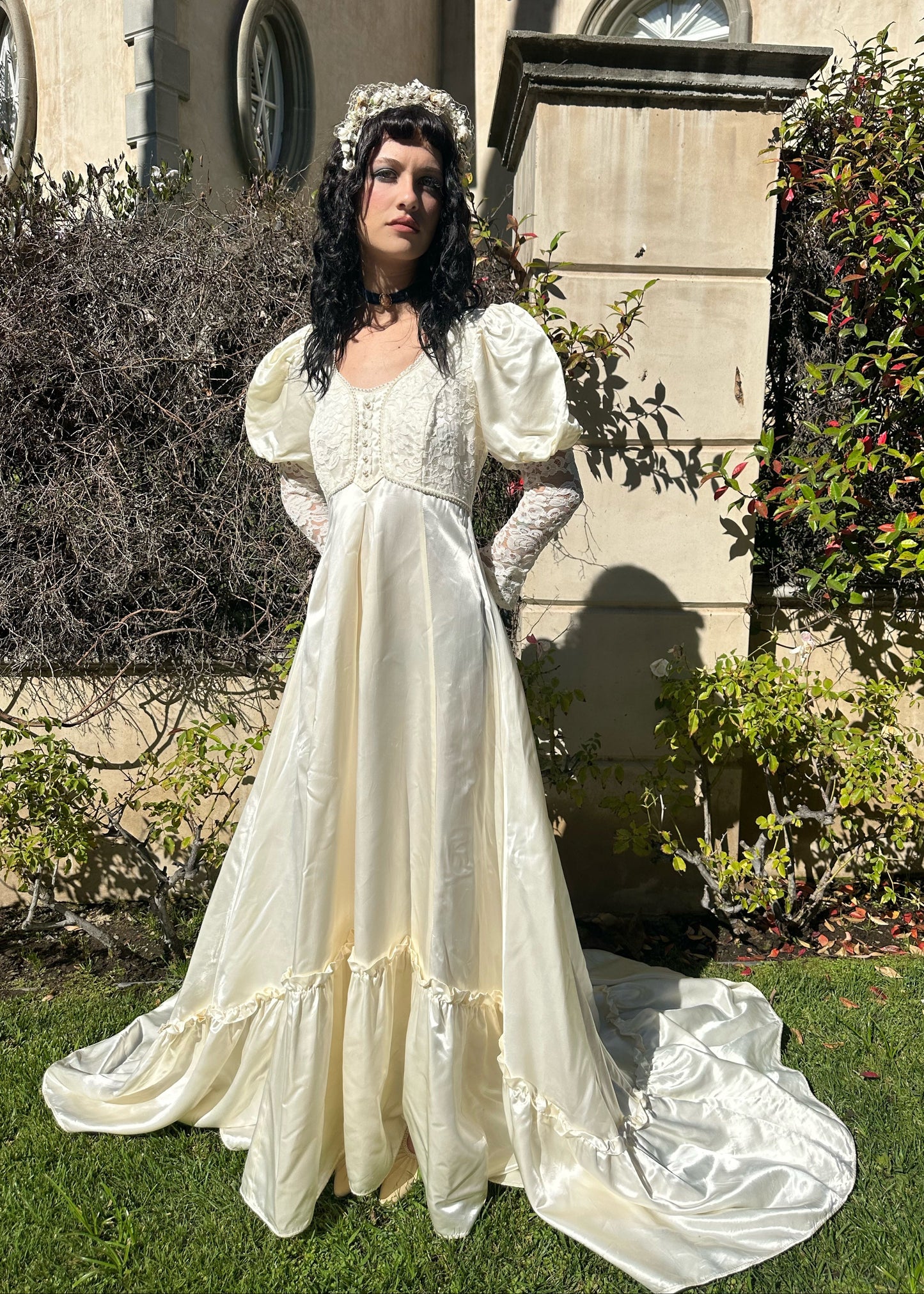 Woman in a white vintage-style dress standing outdoors with a building and greenery in the background