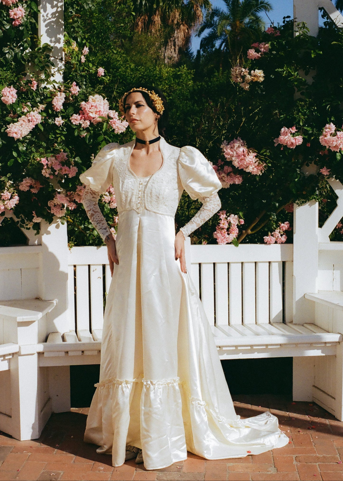 Woman in a white vintage dress standing in front of pink flowers and a white bench.
