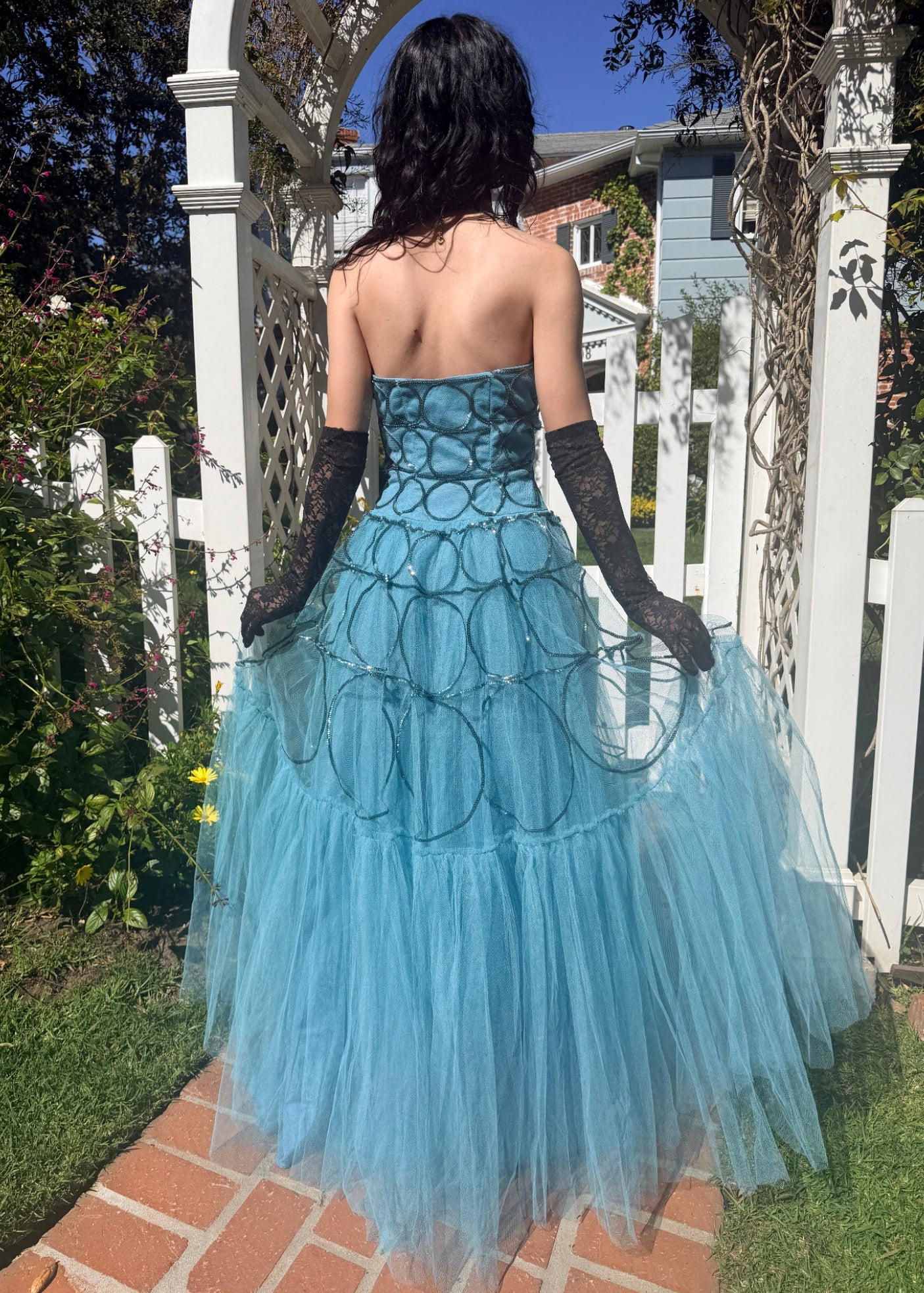 Woman in a 1950's blue strapless ball gown with black gloves standing in a garden.