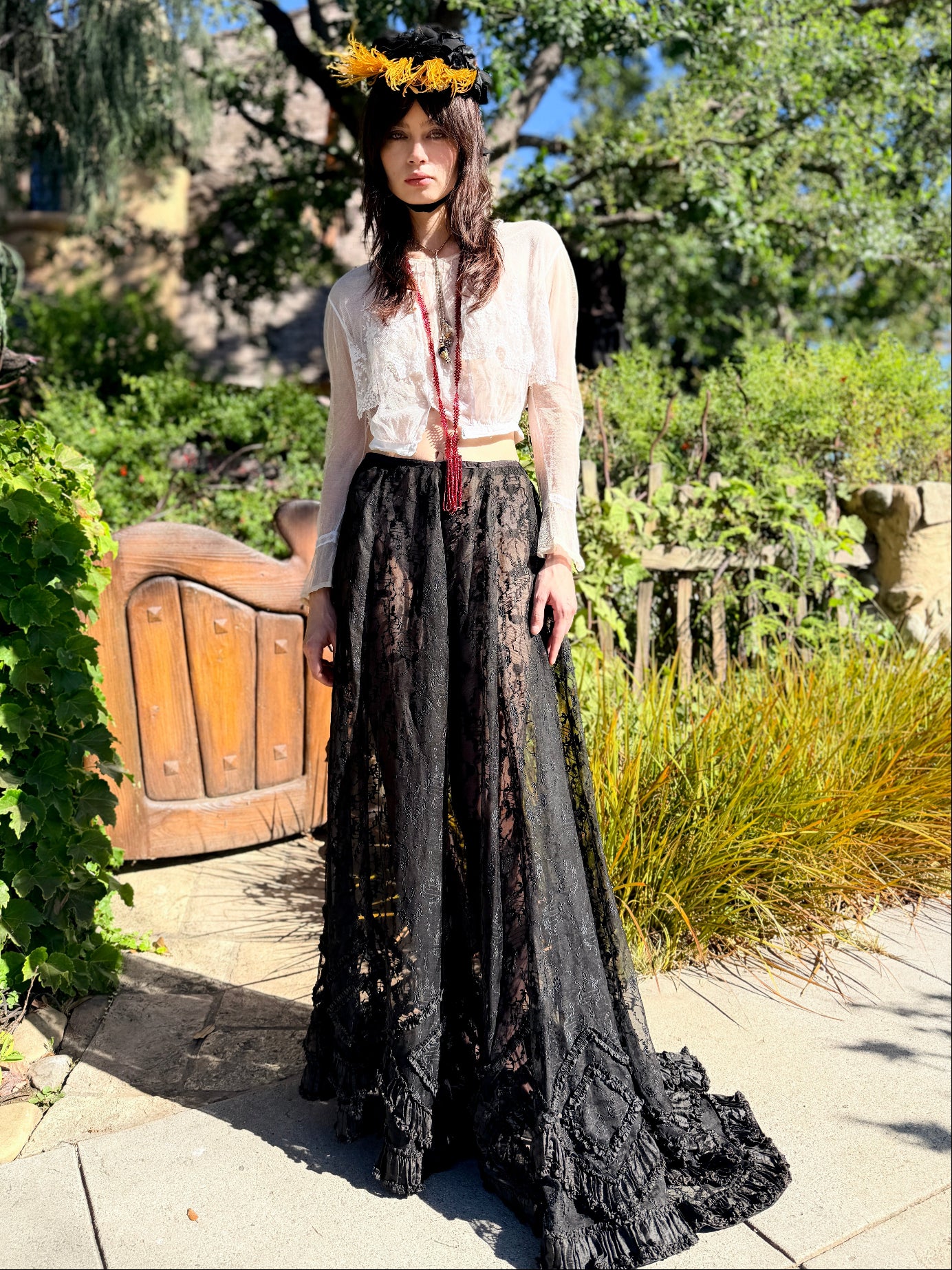 A woman stands outdoors in a garden courtyard wearing a long-sleeve bohemian-style dress with a sheer white lace bodice and a long black floral lace skirt, accessorized with a yellow sunflower crown and a red tassel necklace.
