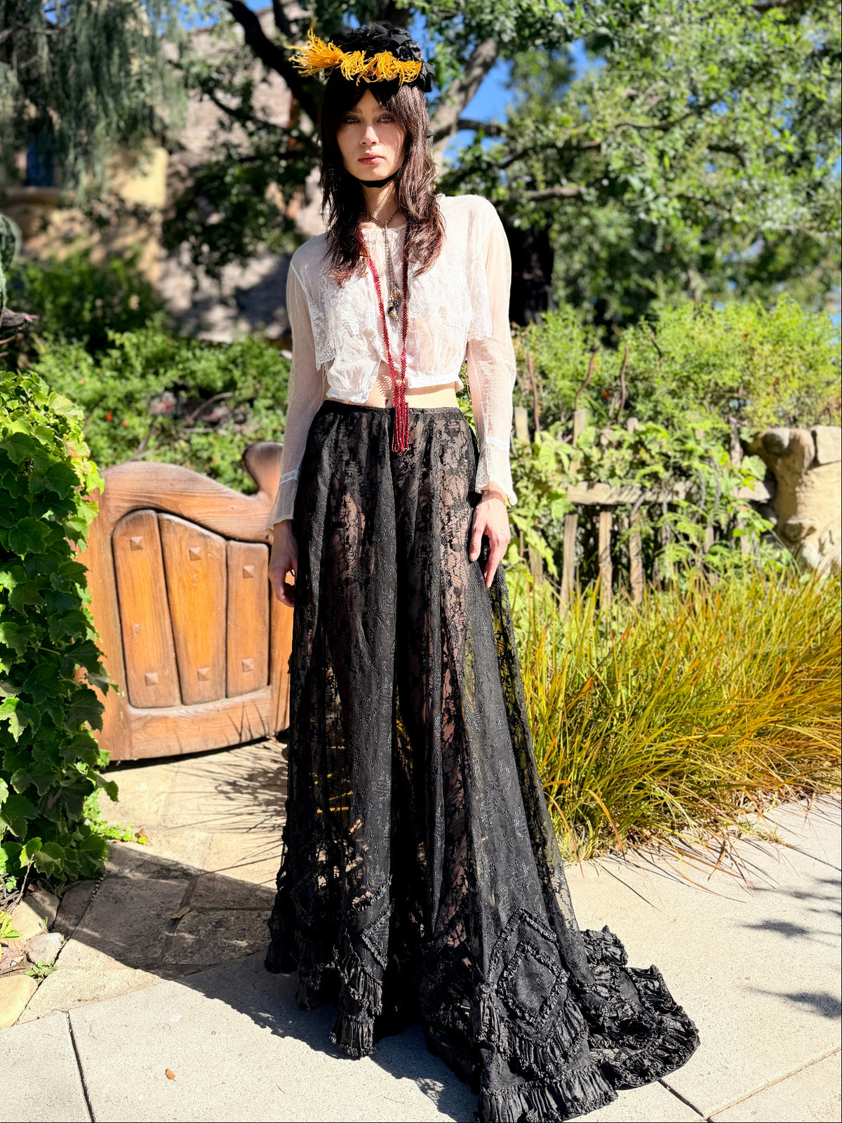 A woman stands outdoors in a garden courtyard wearing a long-sleeve bohemian-style dress with a sheer white lace bodice and a long black floral lace skirt, accessorized with a yellow sunflower crown and a red tassel necklace.