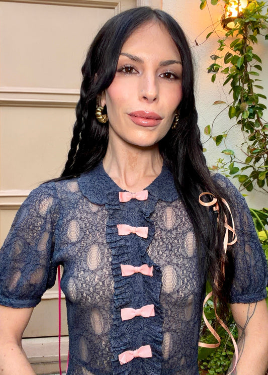 A woman wearing an antique 1930's navy blue lace dress with baby pink bows placed from the neckline all the way to the bottom of the dress, right in the center of the dress. Romantic puff sleeves, bias cut silhouette. 