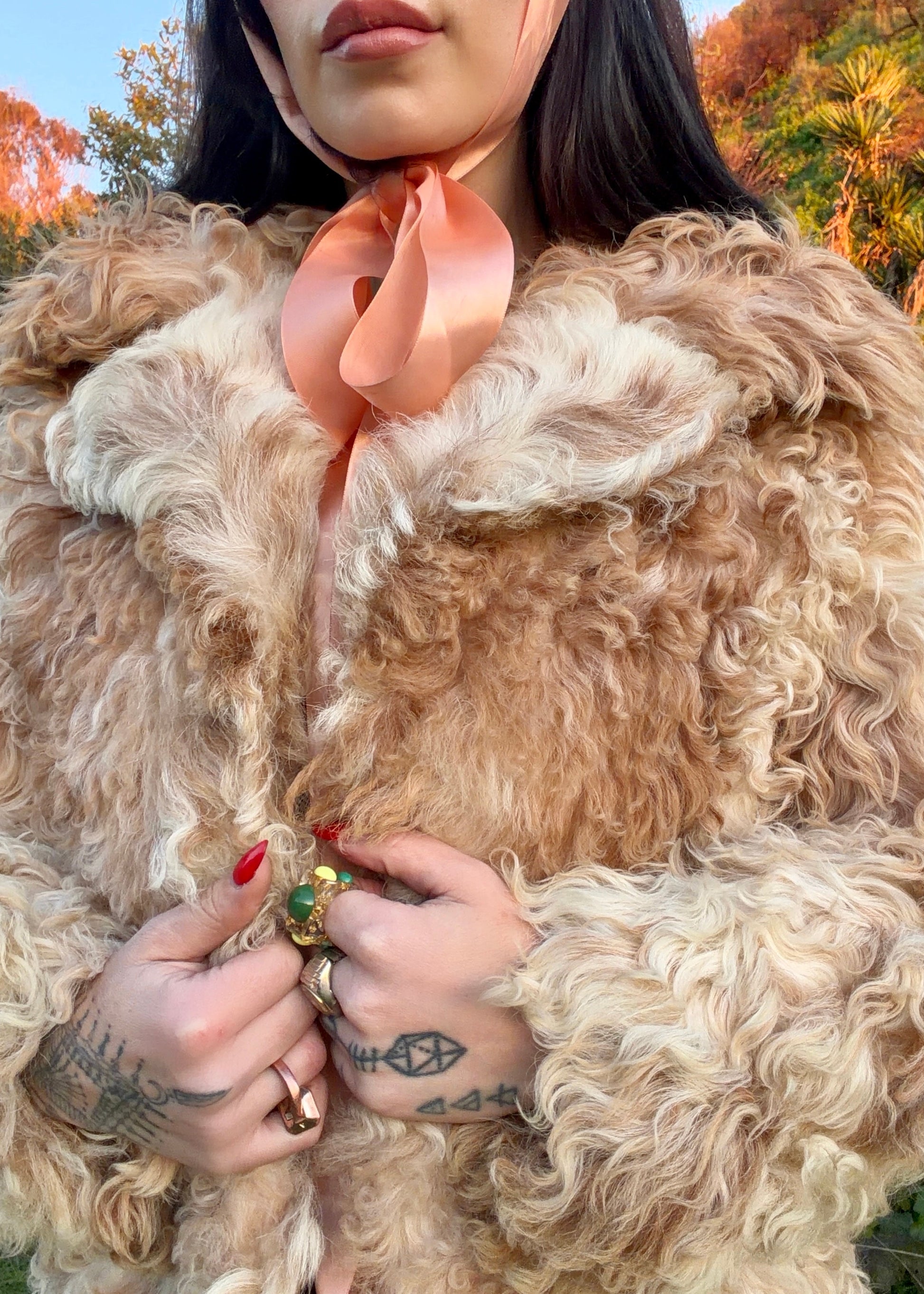 woman wearing a 1970's curly Mongolian and Tibetan sheep's fur coat in fluffy beige and brown colors in a natural setting with trees and grass.