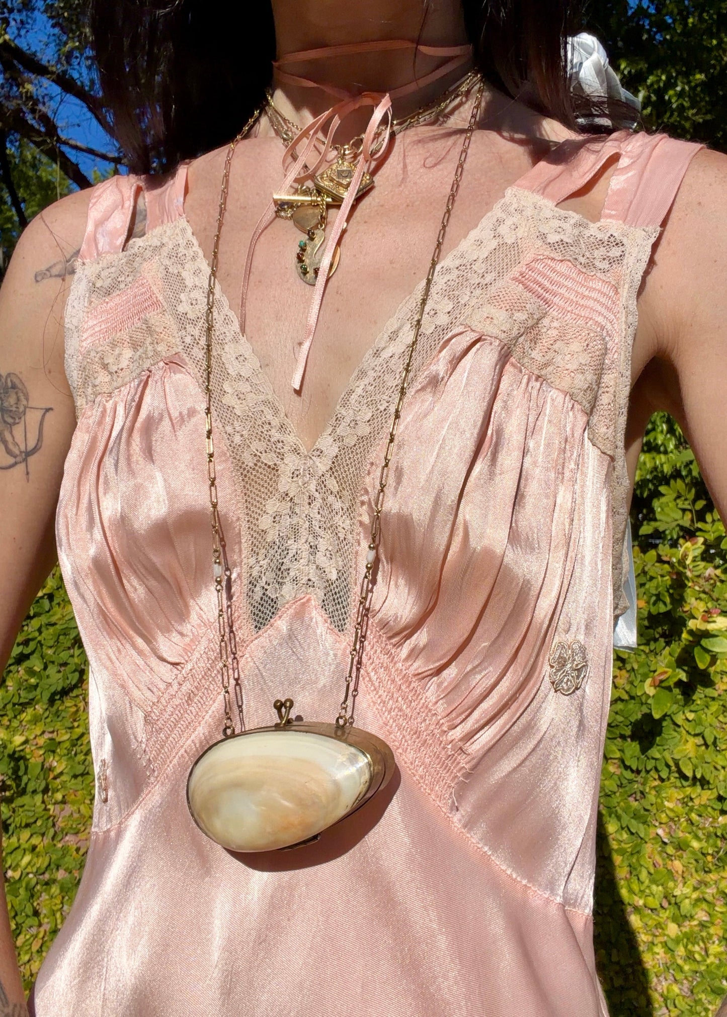 A woman wearing an antique 1930's baby pink silk slip floor-length pale blush satin gown with sheer panels and small floral appliqués, standing outdoors in front of a green hedge and stone path.