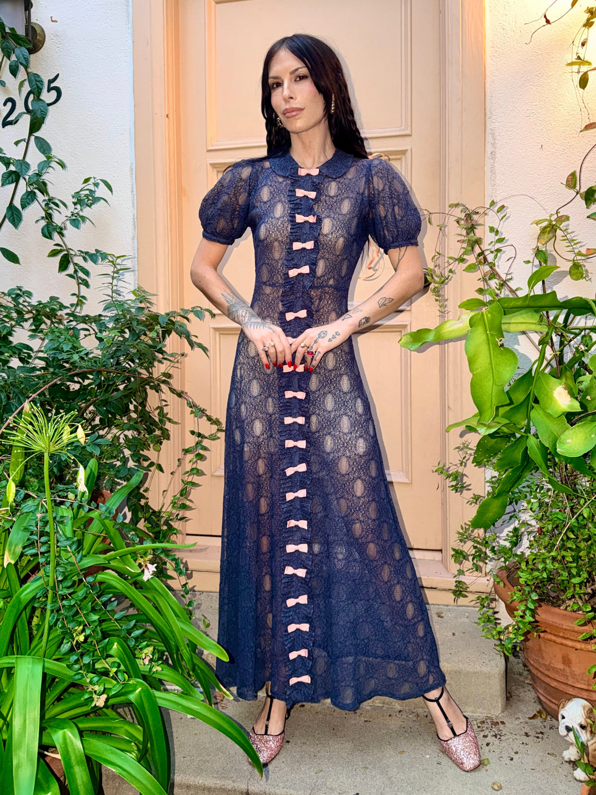 A woman wearing an antique 1930's navy blue lace dress with baby pink bows placed from the neckline all the way to the bottom of the dress, right in the center of the dress. Romantic puff sleeves, bias cut silhouette. 
