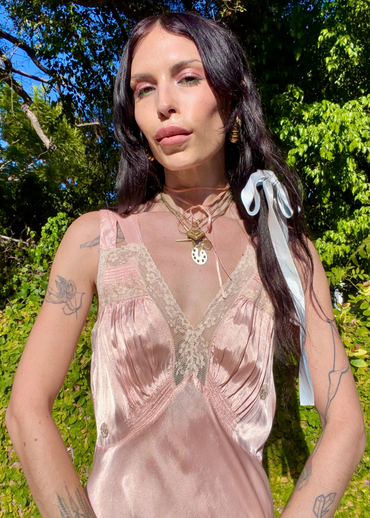 A woman wearing an antique 1930's baby pink silk slip floor-length pale blush satin gown with sheer panels and small floral appliqués, standing outdoors in front of a green hedge and stone path.