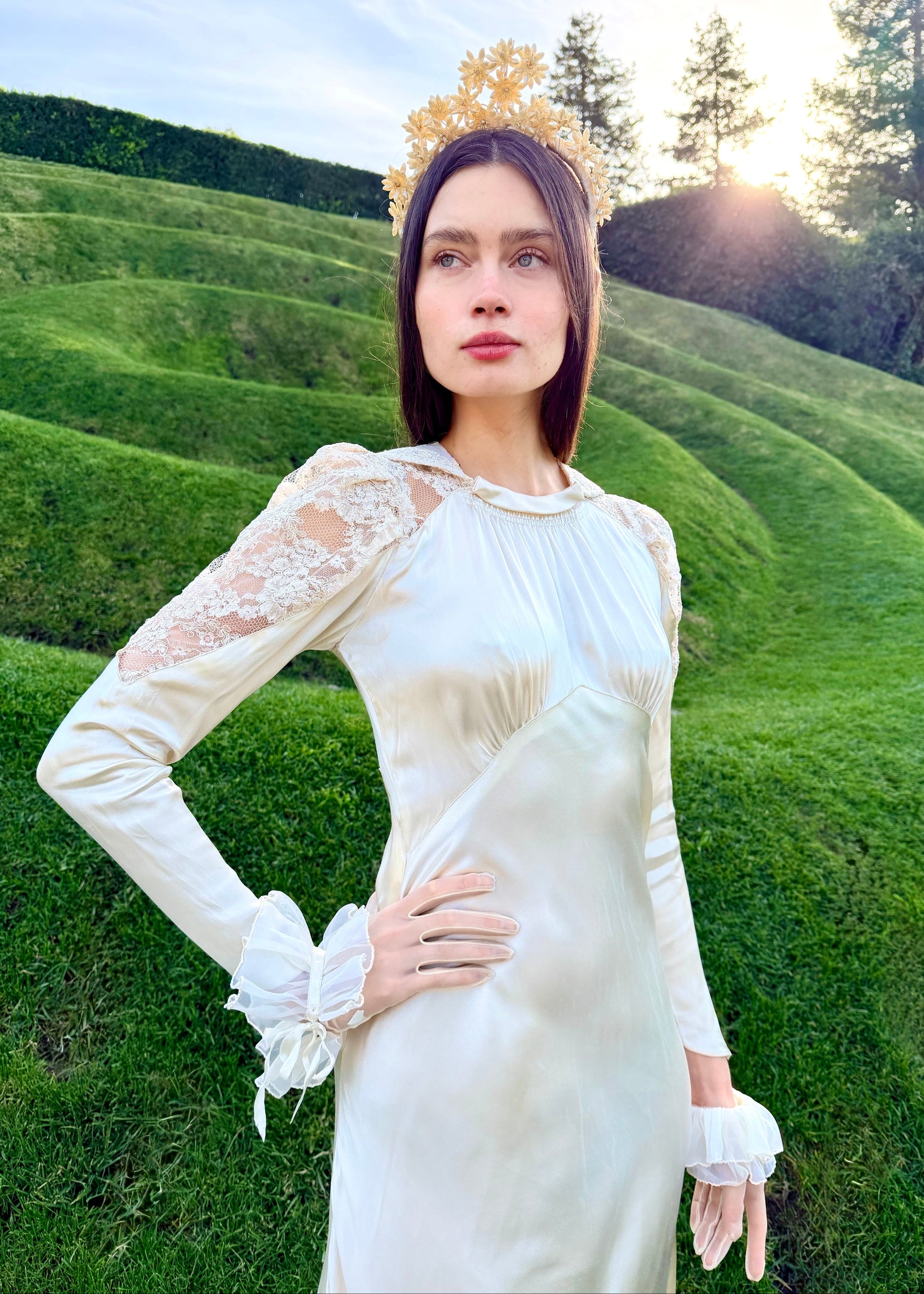 Woman in a white dress with lace sleeves and a gold crown standing in a scenic outdoor setting.