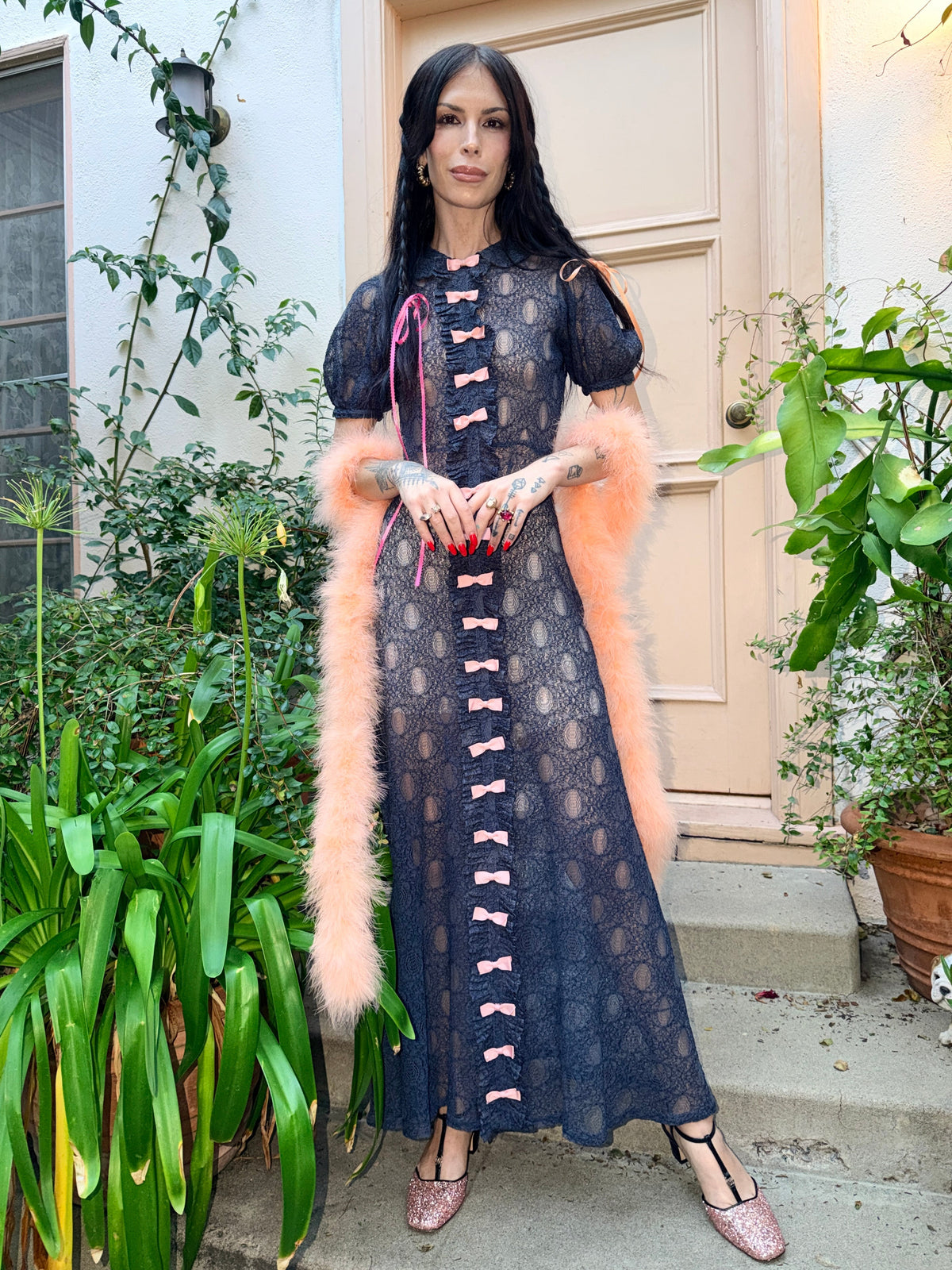 A woman wearing an antique 1930's navy blue lace dress with baby pink bows placed from the neckline all the way to the bottom of the dress, right in the center of the dress. Romantic puff sleeves, bias cut silhouette. 