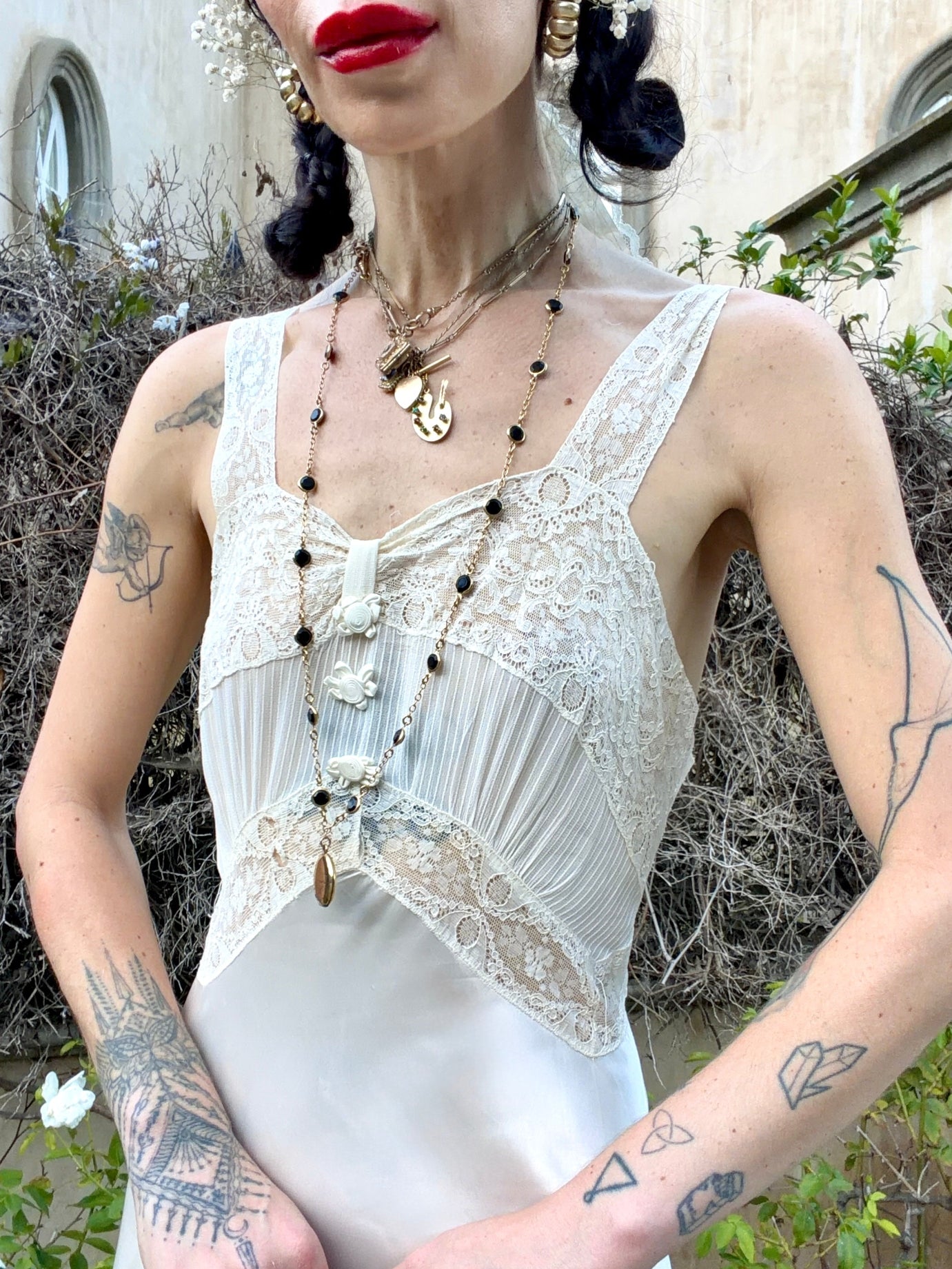 A woman wearing a white lace and satin camisole top with pleated detailing, styled with layered necklaces and earrings, photographed outdoors against a stone wall and greenery.