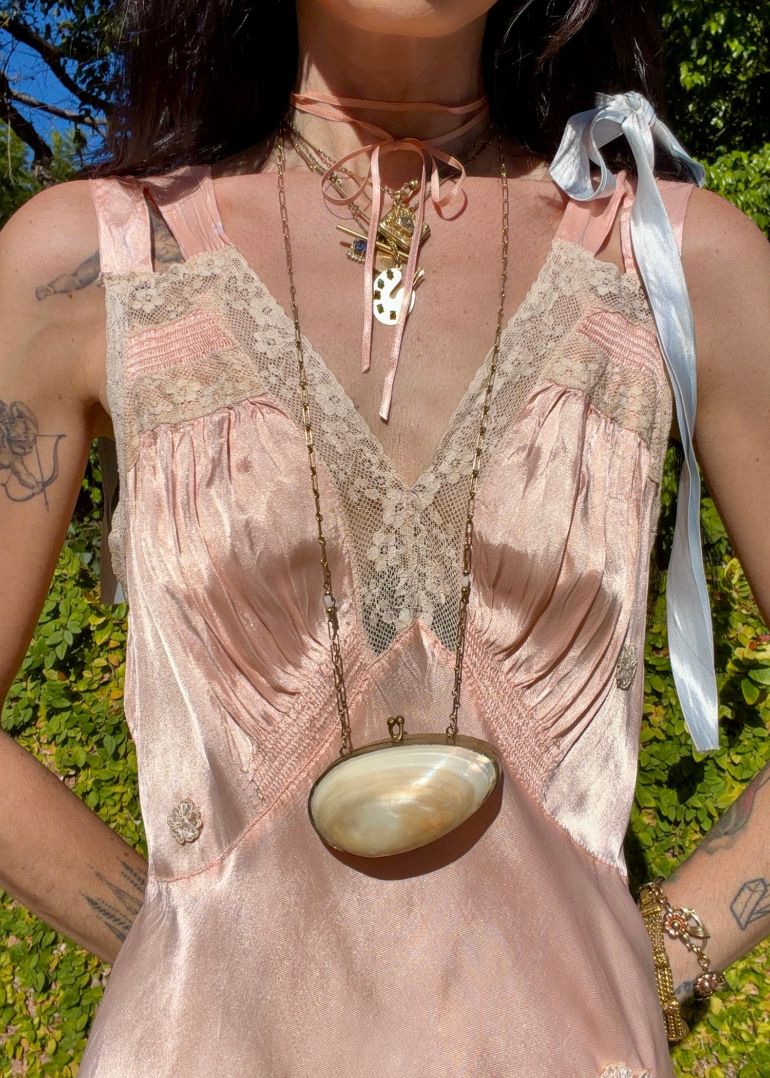 A woman wearing an antique 1930's baby pink silk slip floor-length pale blush satin gown with sheer panels and small floral appliqués, standing outdoors in front of a green hedge and stone path.