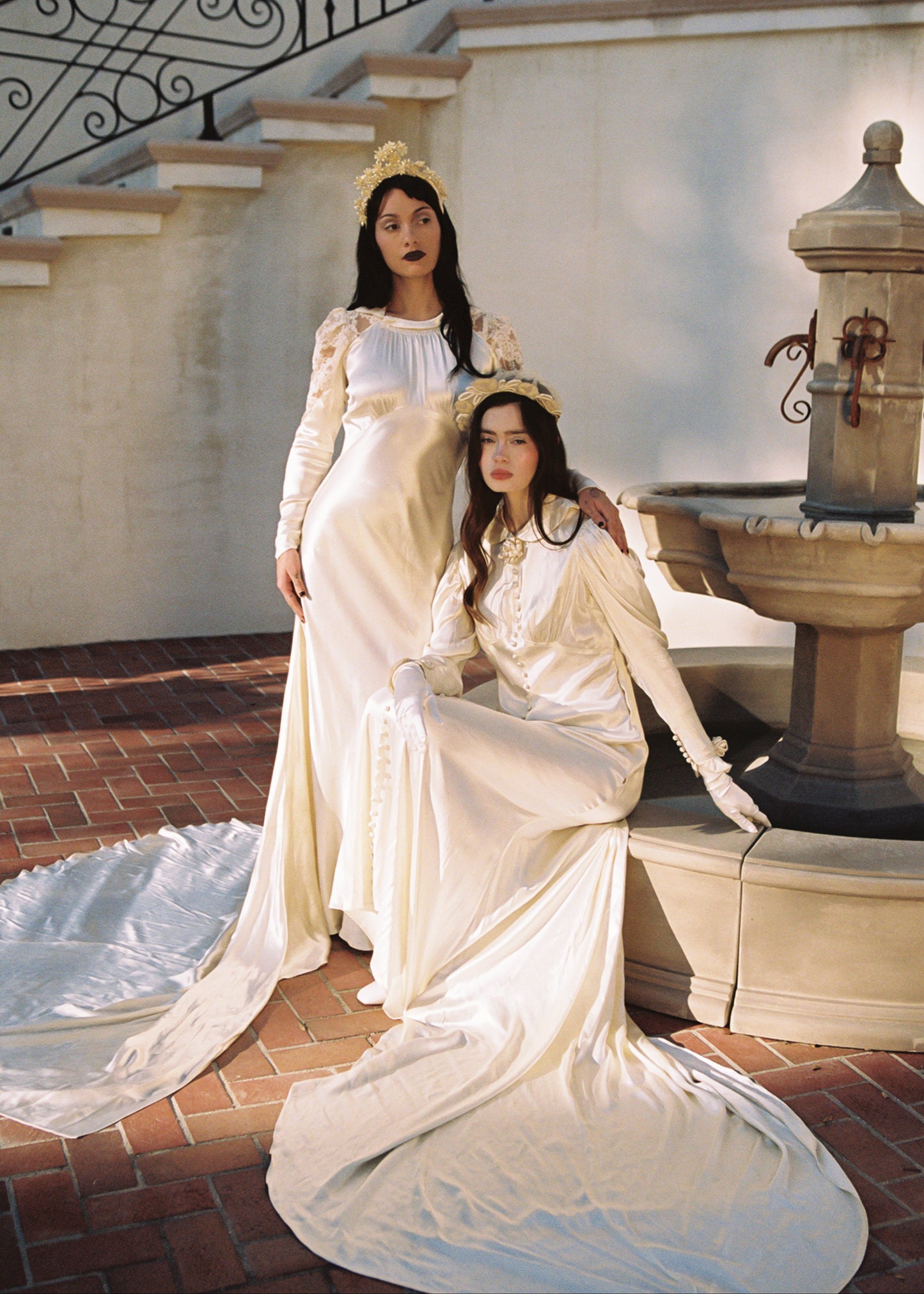 Two women in vintage-style dresses sitting on a stone bench near a fountain.