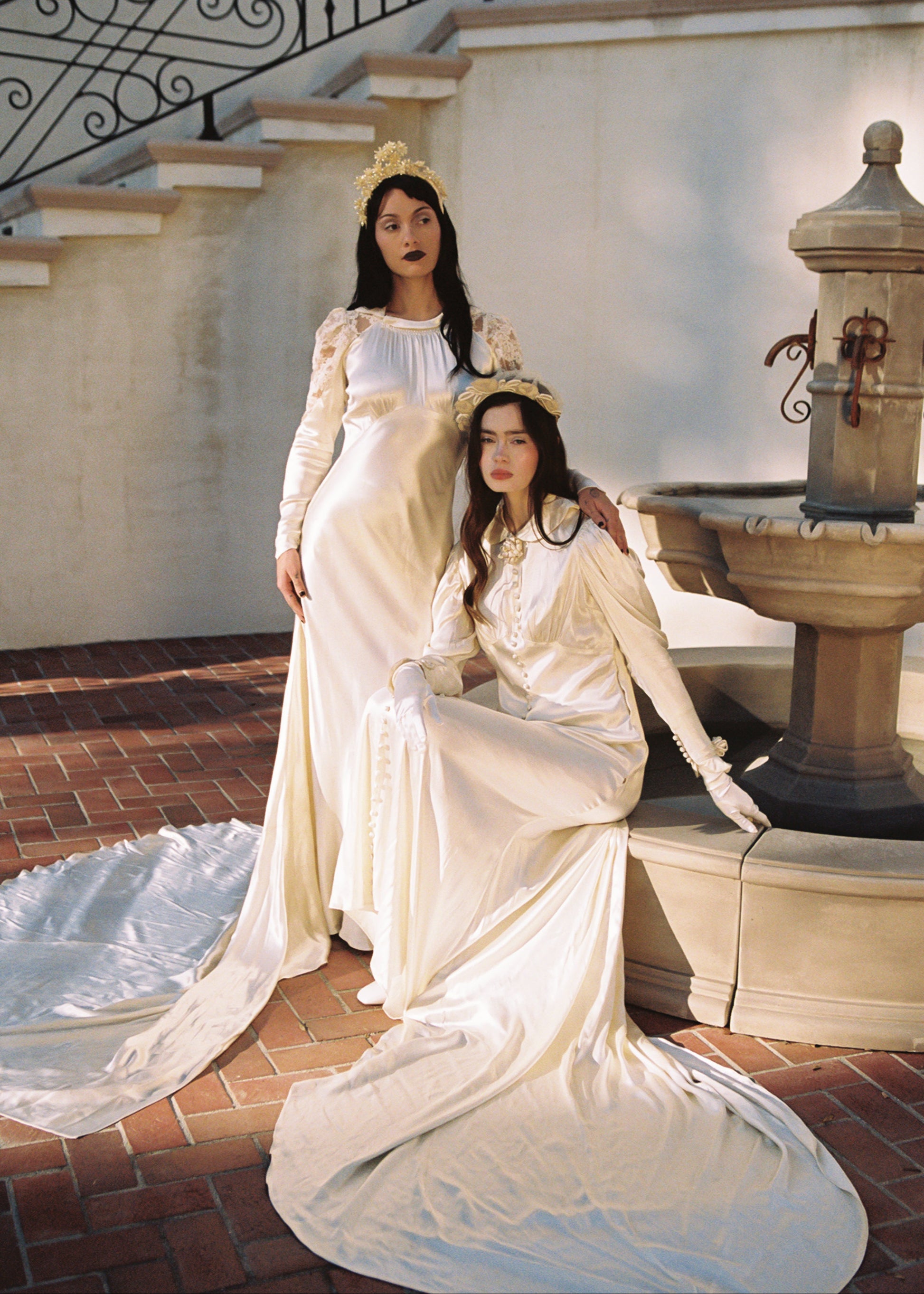 Two women in vintage-style dresses sitting on a stone bench near a fountain.