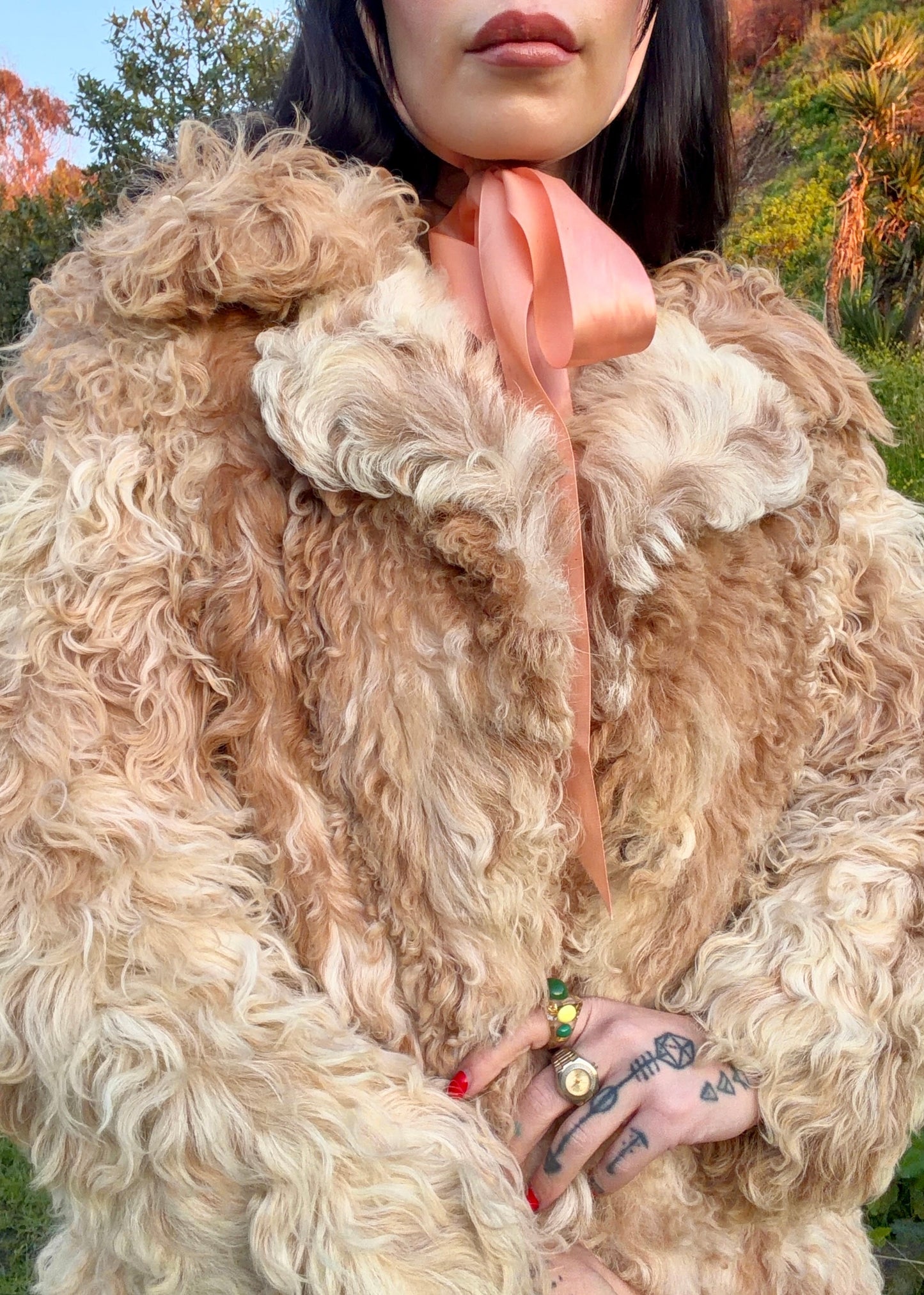 woman wearing a 1970's curly Mongolian and Tibetan sheep's fur coat in fluffy beige and brown colors in a natural setting with trees and grass.