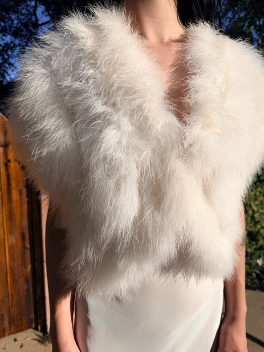 Person wearing a white marabou vest outdoors with trees and a wooden fence in the background