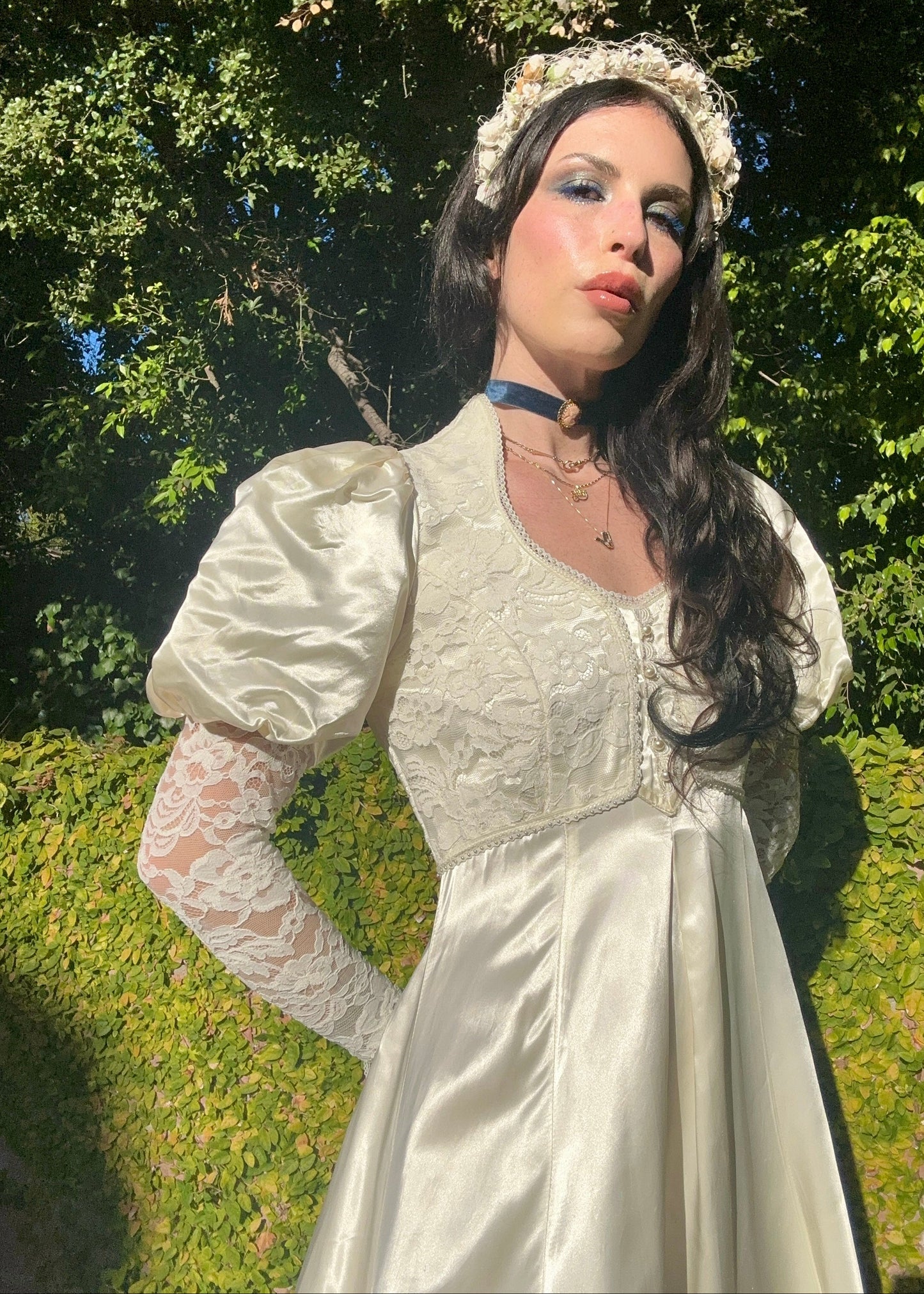 Woman in a Gunne Sax vintage dress with lace sleeves and a headband, standing outdoors with greenery in the background.