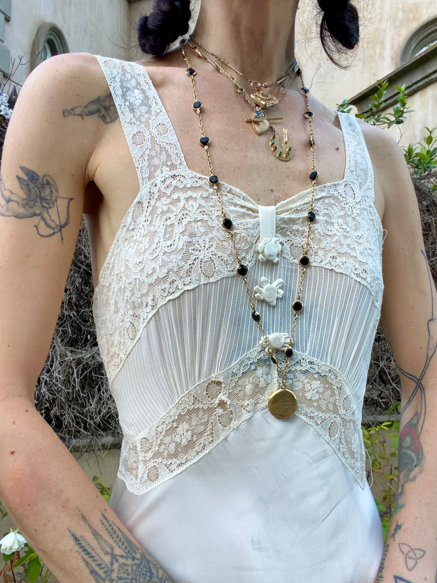 A close-up of a person wearing a 1930s silk slip white lace and pleated camisole-style top with layered gold and black beaded necklaces, photographed outdoors against a stone wall and greenery.