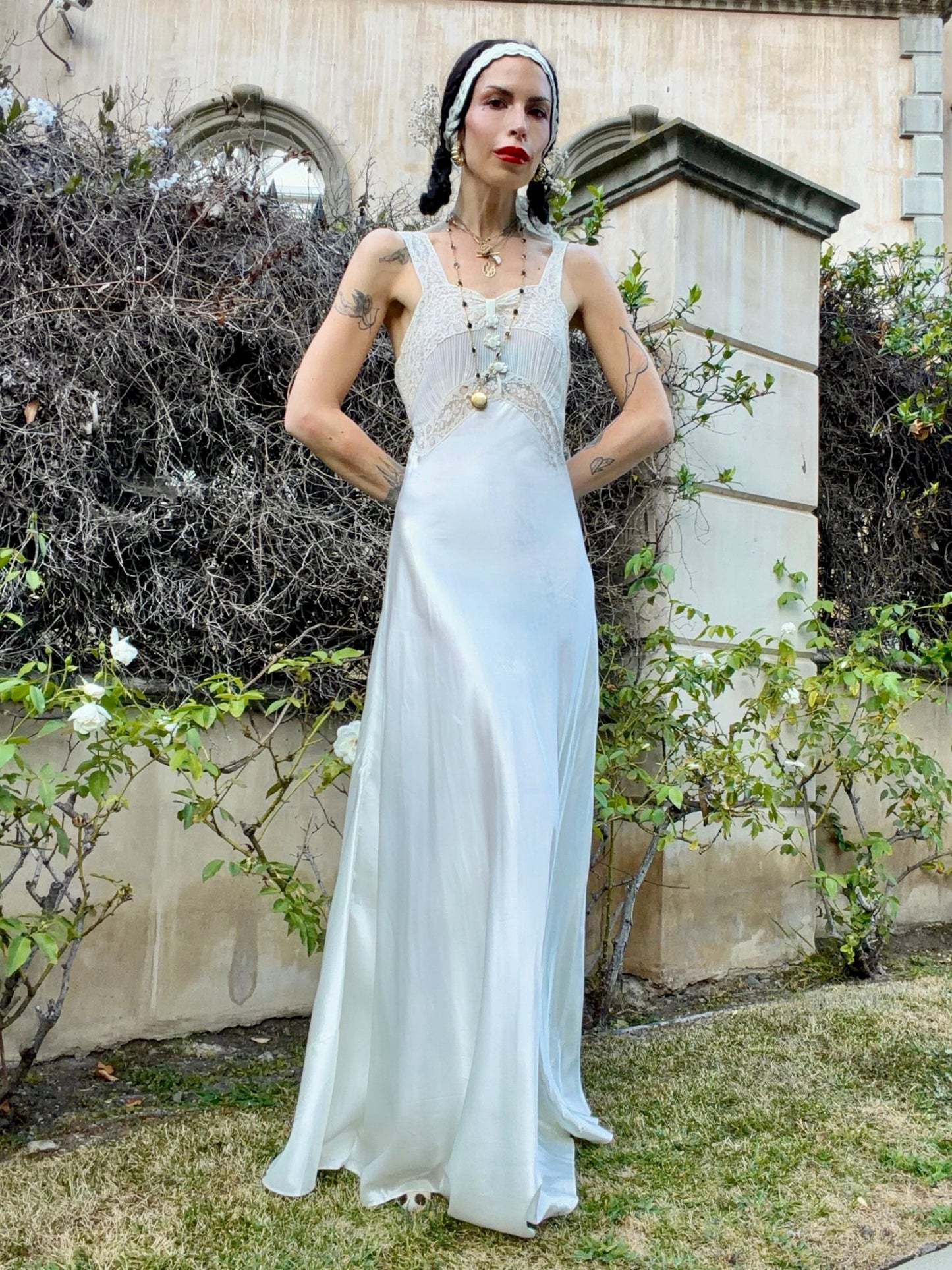 A woman stands outdoors in front of a stone wall and greenery, wearing a sleeveless white satin gown with a beaded bodice, layered necklaces, and a headpiece.