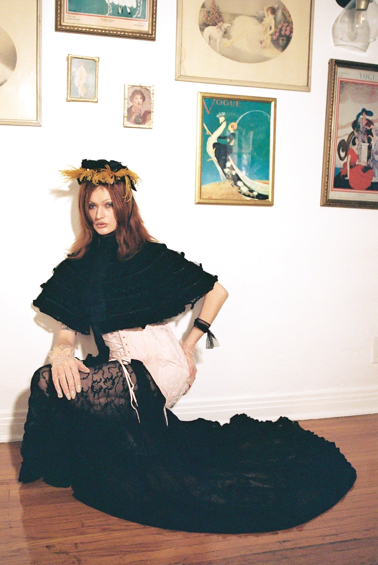 A woman kneels on a wooden floor against a white wall covered with framed vintage art prints. She is wearing a black ruffled cape, a pale pink corset-style top, a black lace skirt, and a black headpiece with yellow flowers.