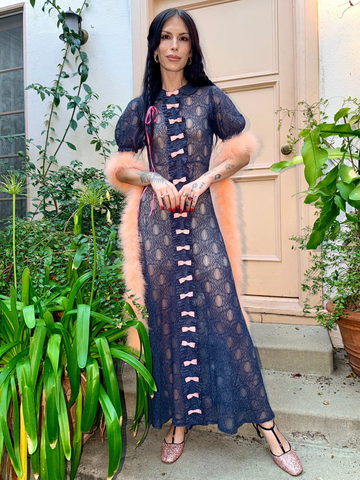 A woman wearing an antique 1930's navy blue lace dress with baby pink bows placed from the neckline all the way to the bottom of the dress, right in the center of the dress. Romantic puff sleeves, bias cut silhouette. 