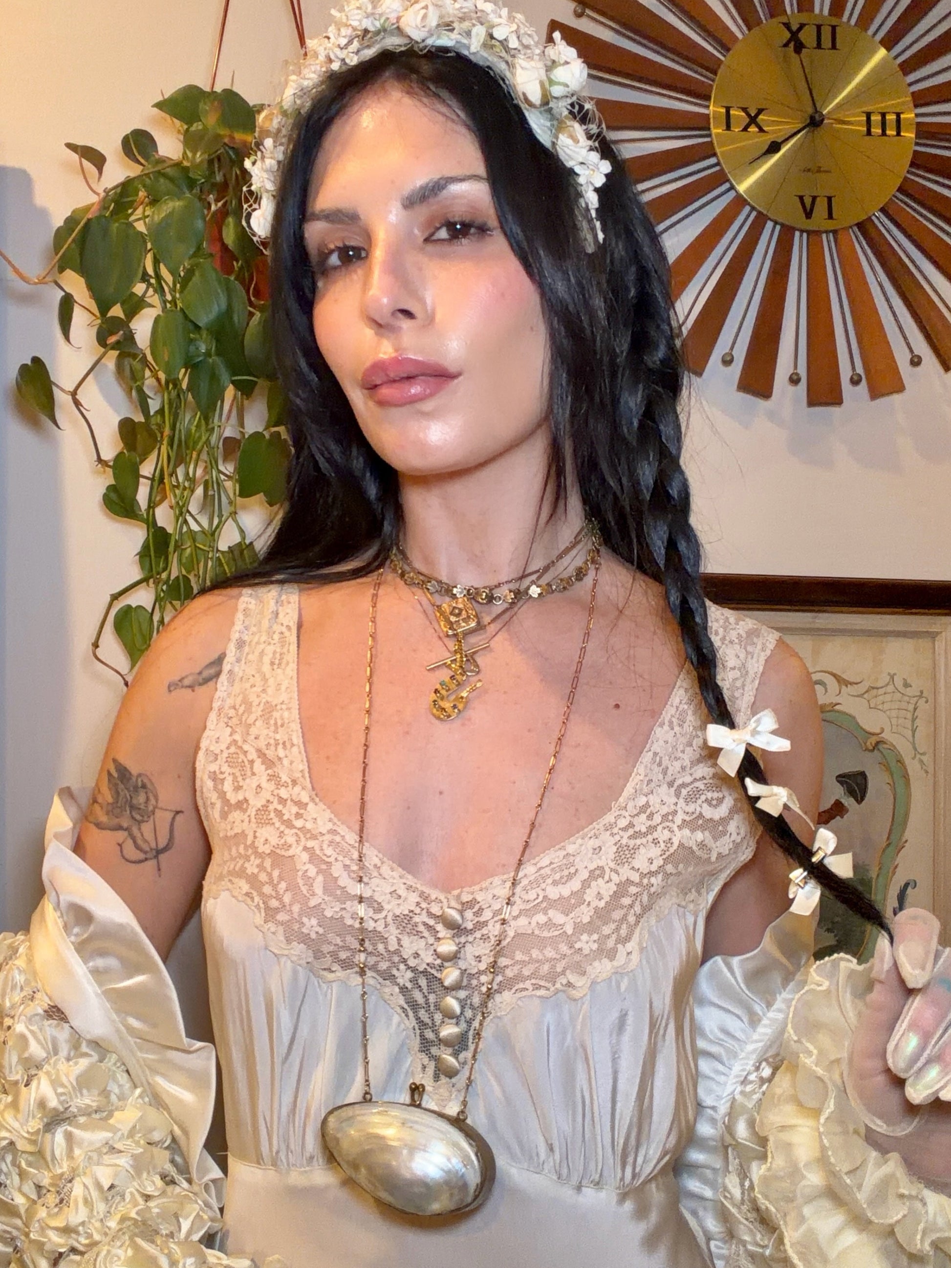 Woman in a white dress with decorative headpiece and jewelry, holding a spoon, in a room with a clock and plant.