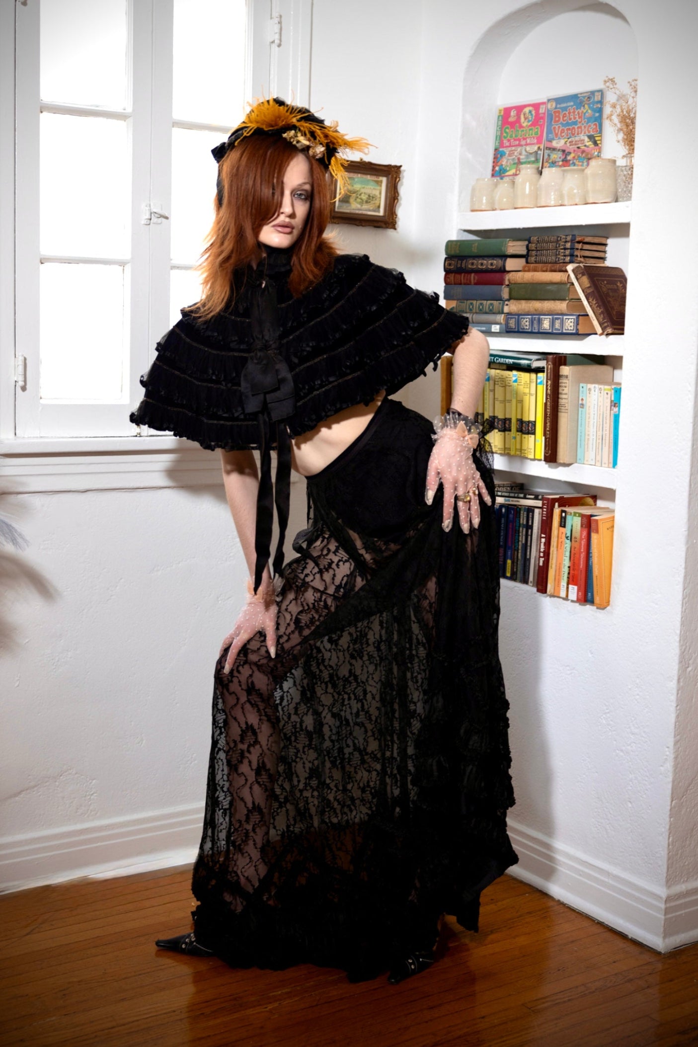 A woman with red hair wearing a black Victorian-style lace dress and a black ruffled cape with a yellow floral headpiece, posing in a room with white walls, a window, and a bookshelf.