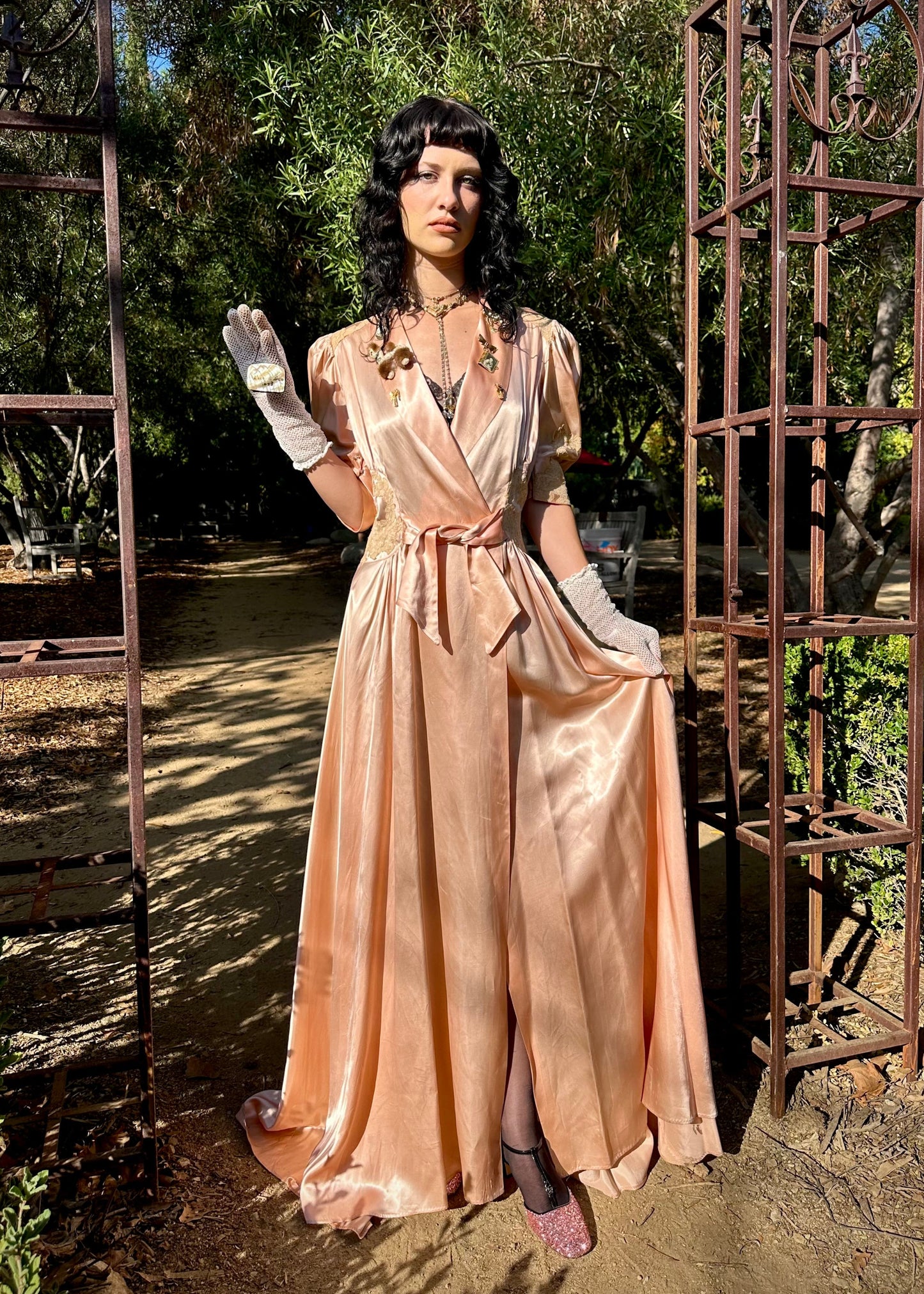 Person in a vintage-style peach dress standing outdoors with metal arches and greenery.