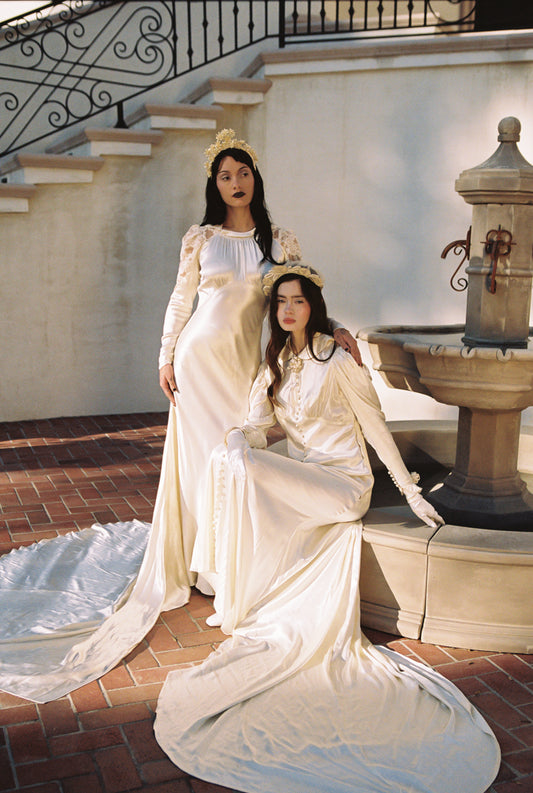 Two women in antique 1930's art deco style wedding dresses sitting on a stone bench near a fountain.