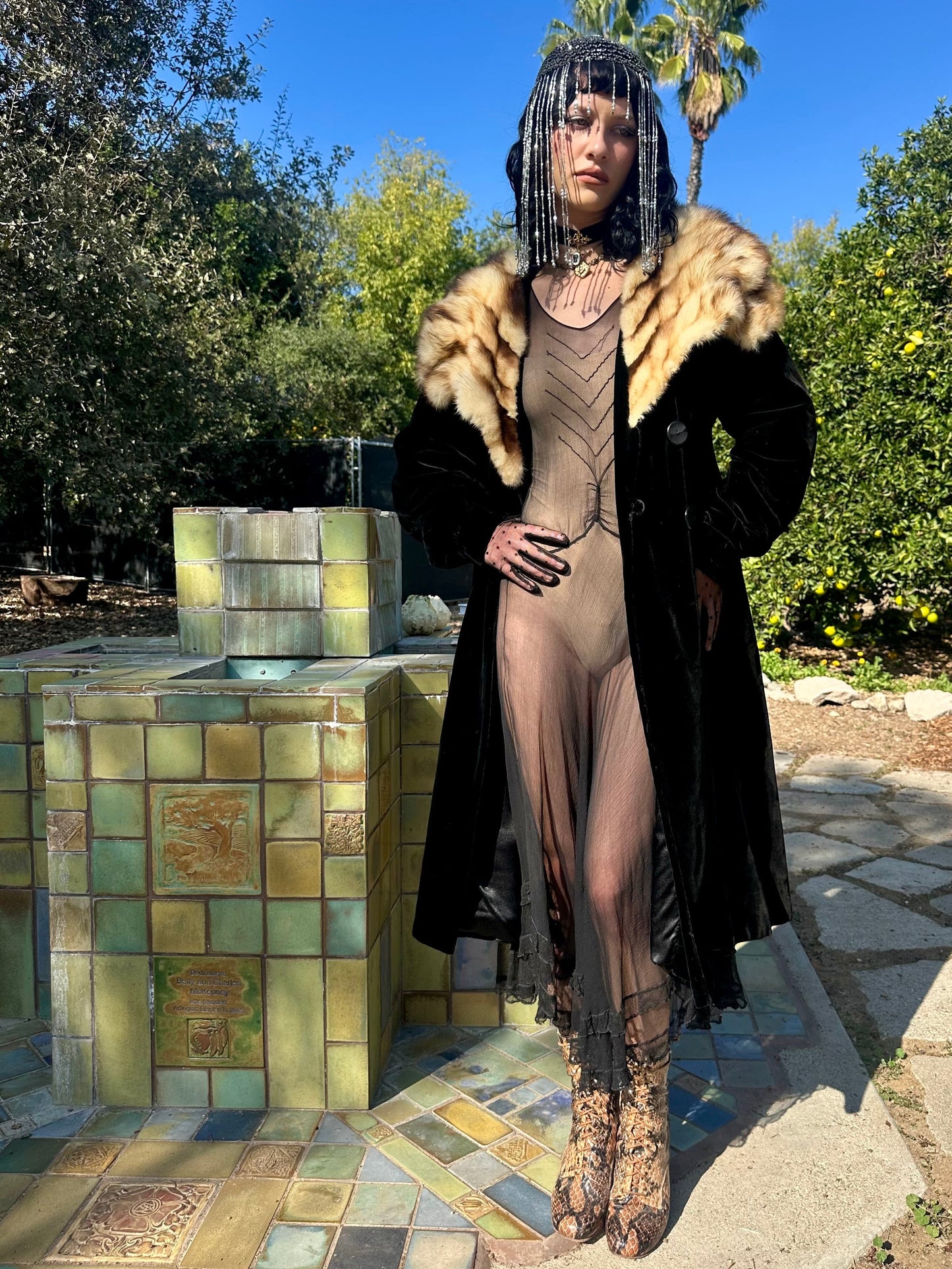 Person in an antique 1930s opera coat standing outdoors with trees and a fountain in the background
