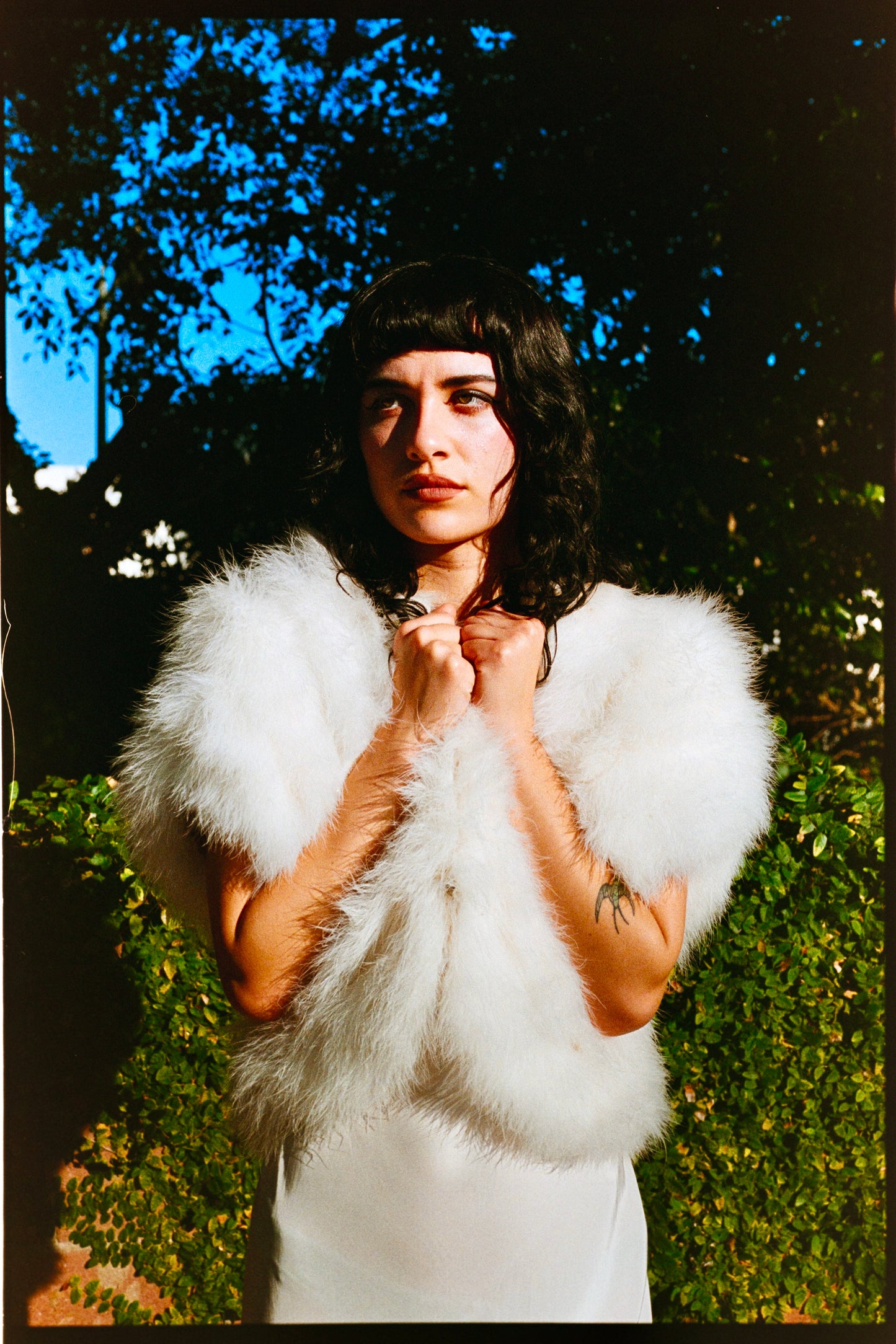 Person wearing a white marabou vest outdoors with trees and a wooden fence in the background