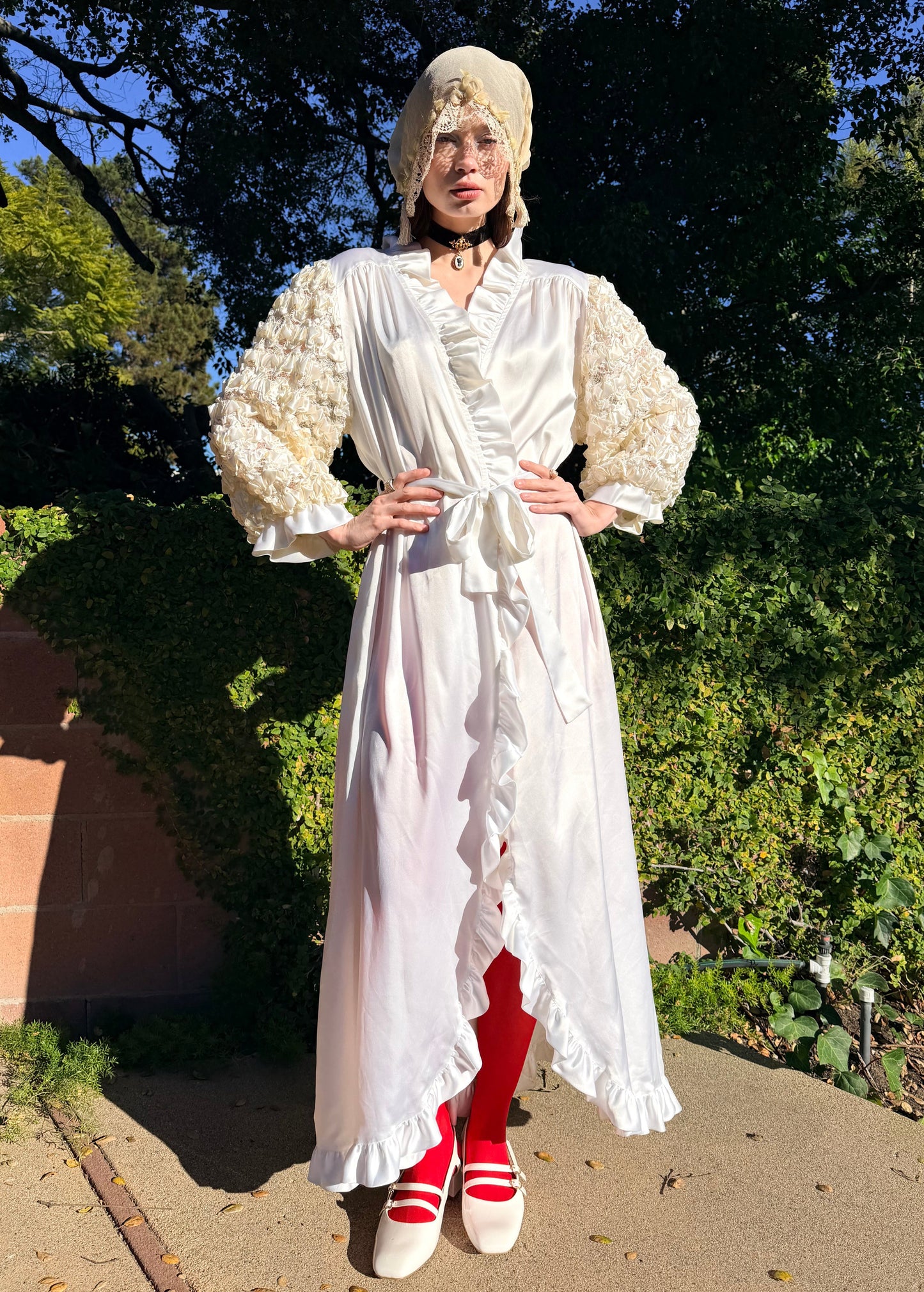 Person wearing a white dress with red shoes standing outdoors with trees in the background