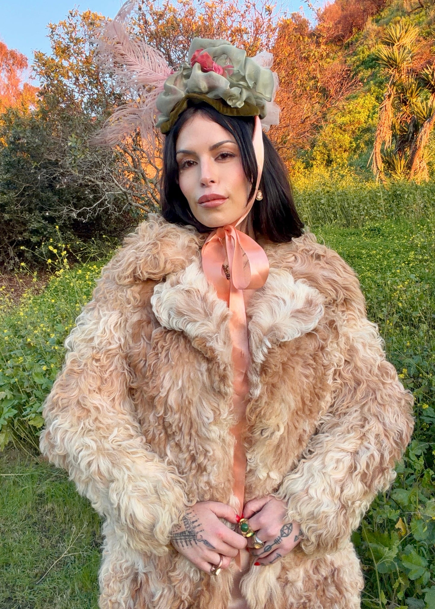 woman wearing a 1970's curly Mongolian and Tibetan sheep's fur coat in fluffy beige and brown colors in a natural setting with trees and grass.