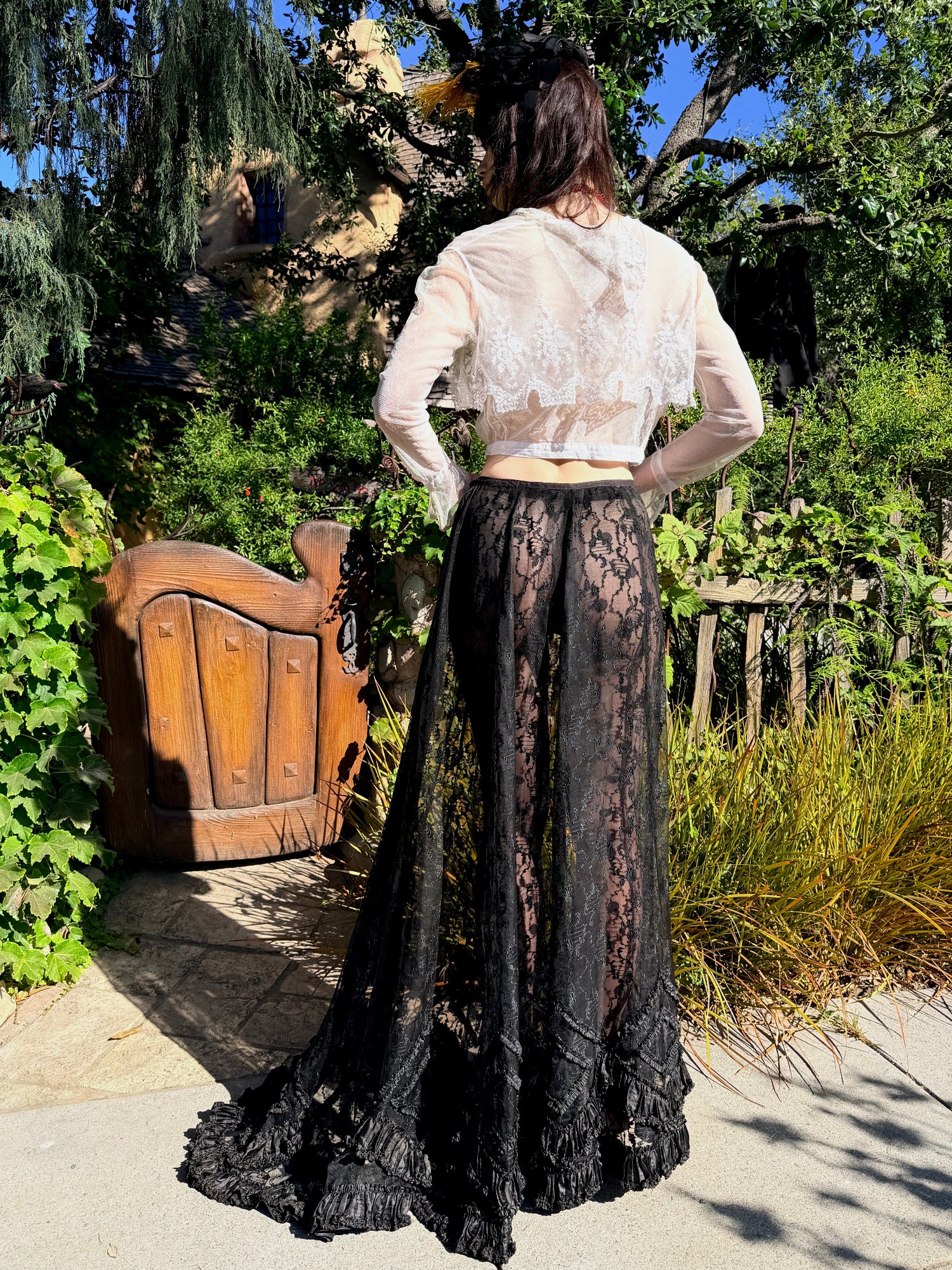 A woman stands outdoors in a garden setting, viewed from behind, wearing a long black lace skirt with a sheer cream lace cropped top.
