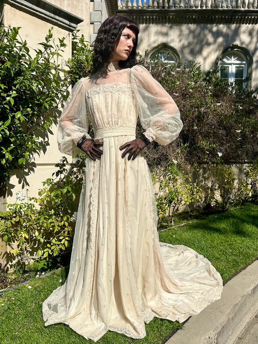 Woman in a long, white lace dress standing outdoors with greenery and a building in the background.