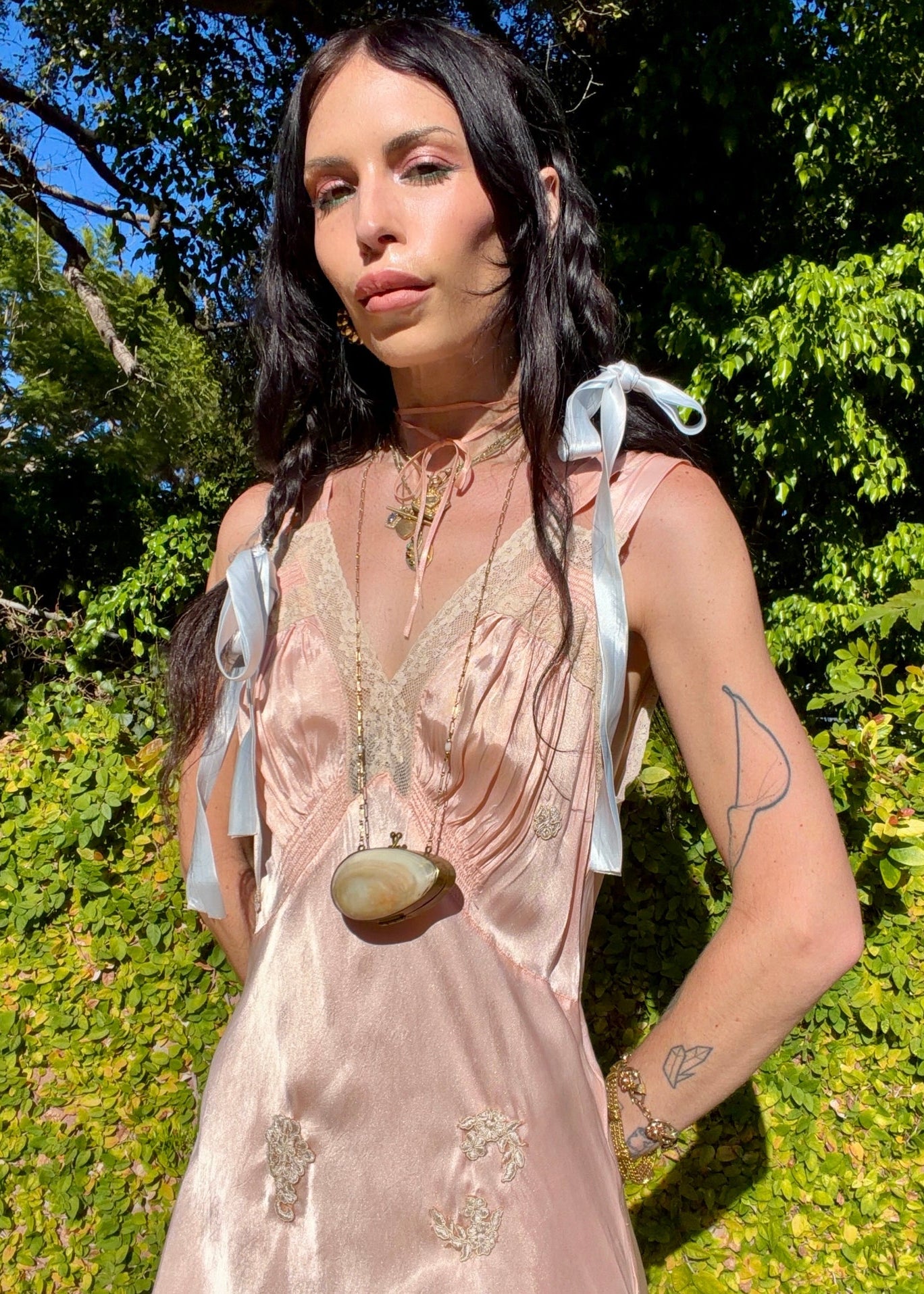A woman wearing an antique 1930's baby pink silk slip floor-length pale blush satin gown with sheer panels and small floral appliqués, standing outdoors in front of a green hedge and stone path.