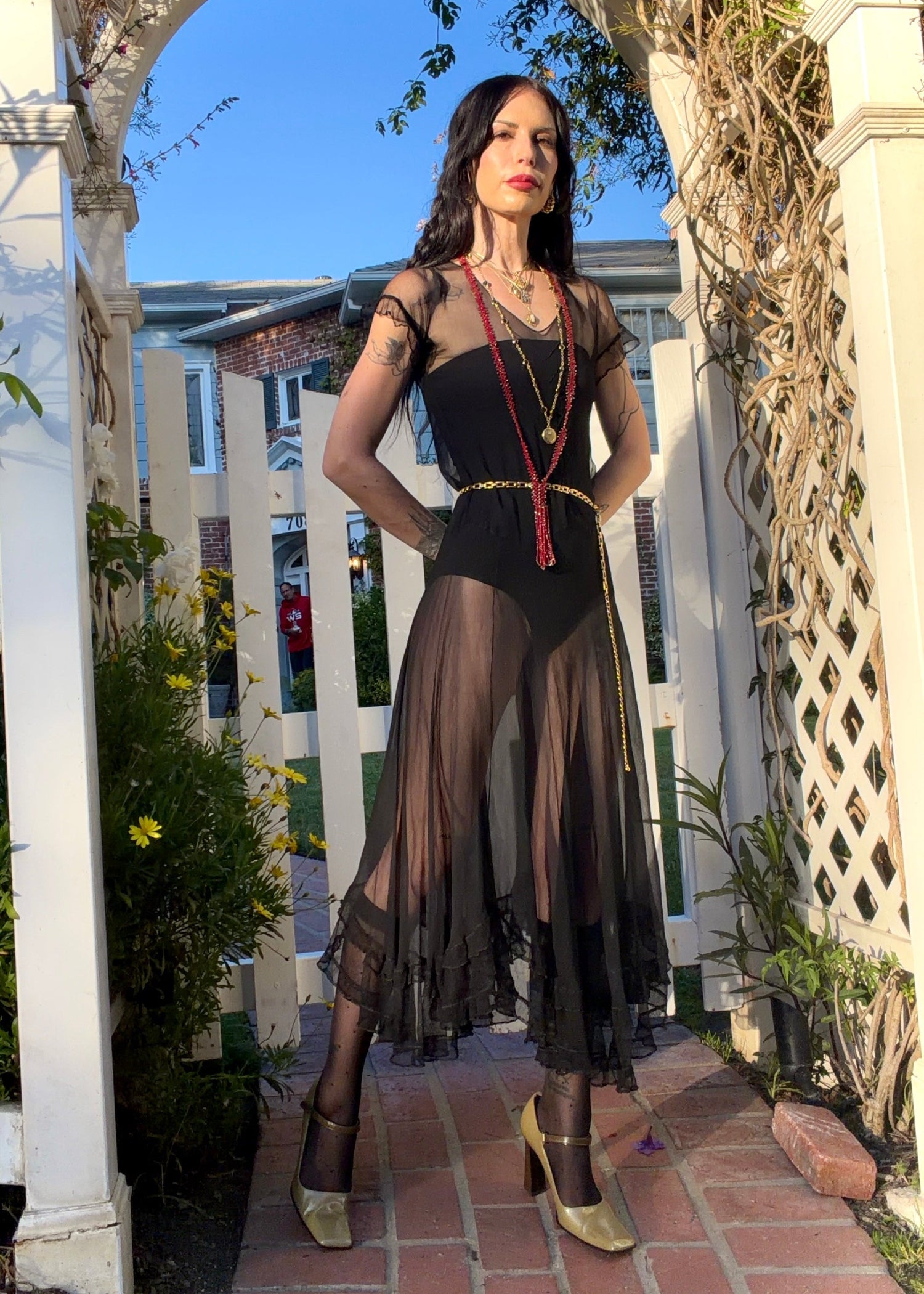 A woman stands outdoors under a white arched garden gate wearing a sheer black antique 1920's dress with a long black mesh skirt, layered gold necklaces, a gold chain belt, and beige high heels.