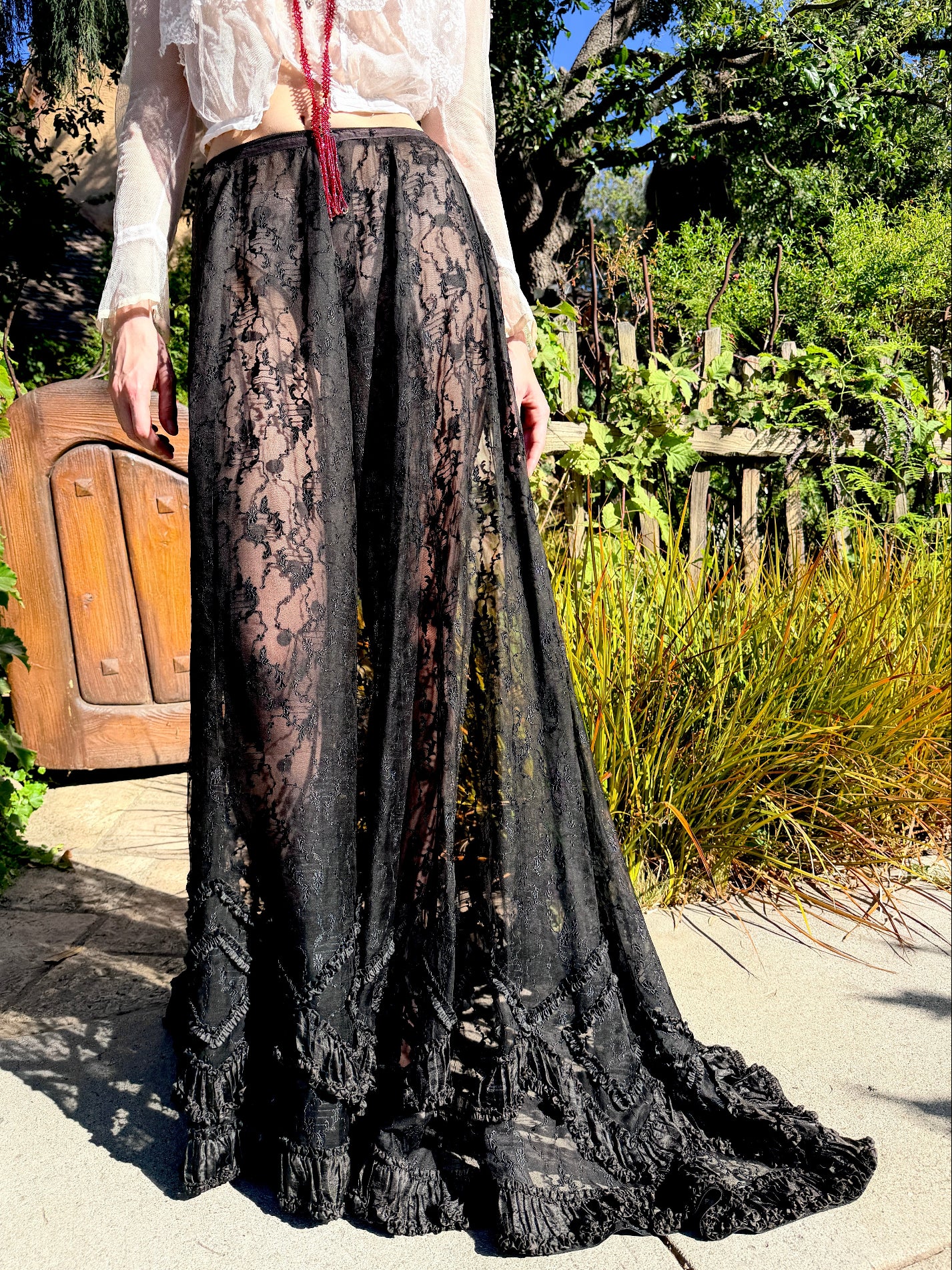 A person outdoors wearing a long black lace skirt with a dramatic train, paired with a white sheer long-sleeve top and a red tassel necklace. The setting is a sunny garden with green foliage, a wooden fence, and a stone path.