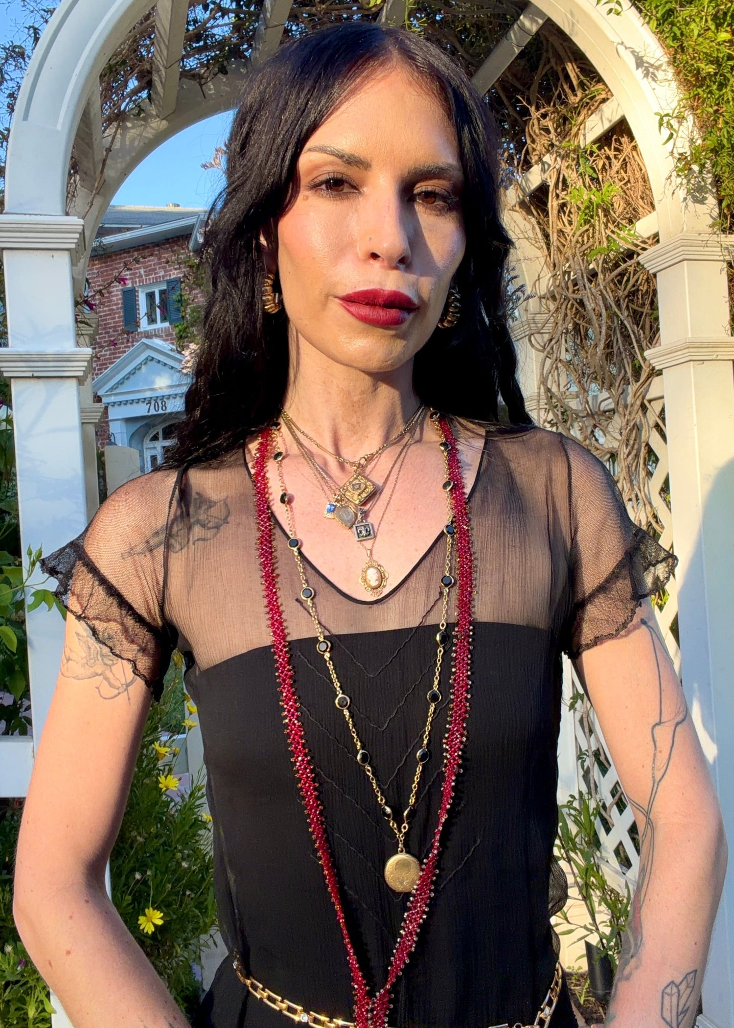 A woman stands outdoors under a white arched garden gate wearing a sheer black antique 1920's dress with a long black mesh skirt, layered gold necklaces, a gold chain belt, and beige high heels.