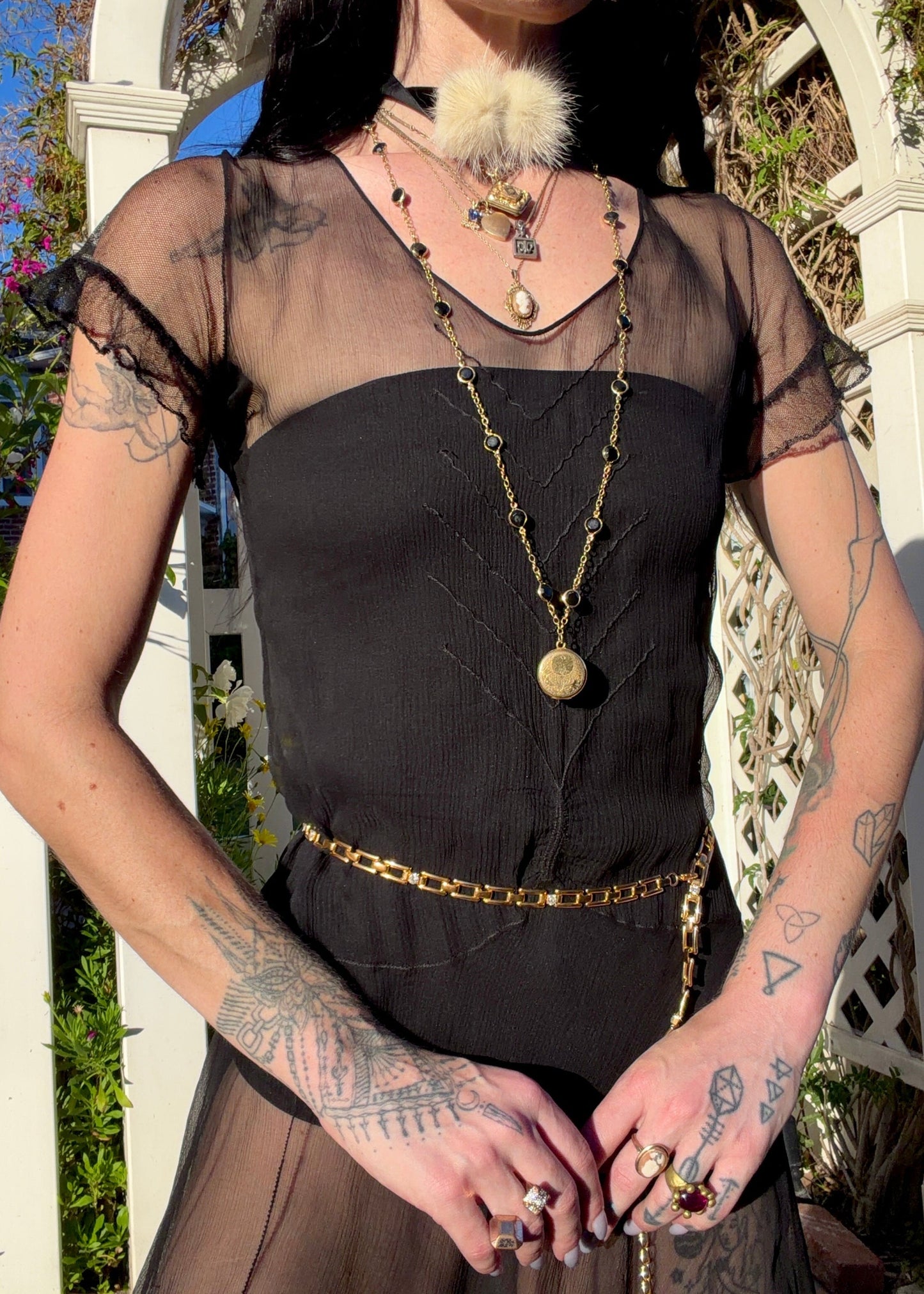 A woman stands outdoors under a white arched garden gate wearing a sheer black antique 1920's dress with a long black mesh skirt, layered gold necklaces, a gold chain belt, and beige high heels.
