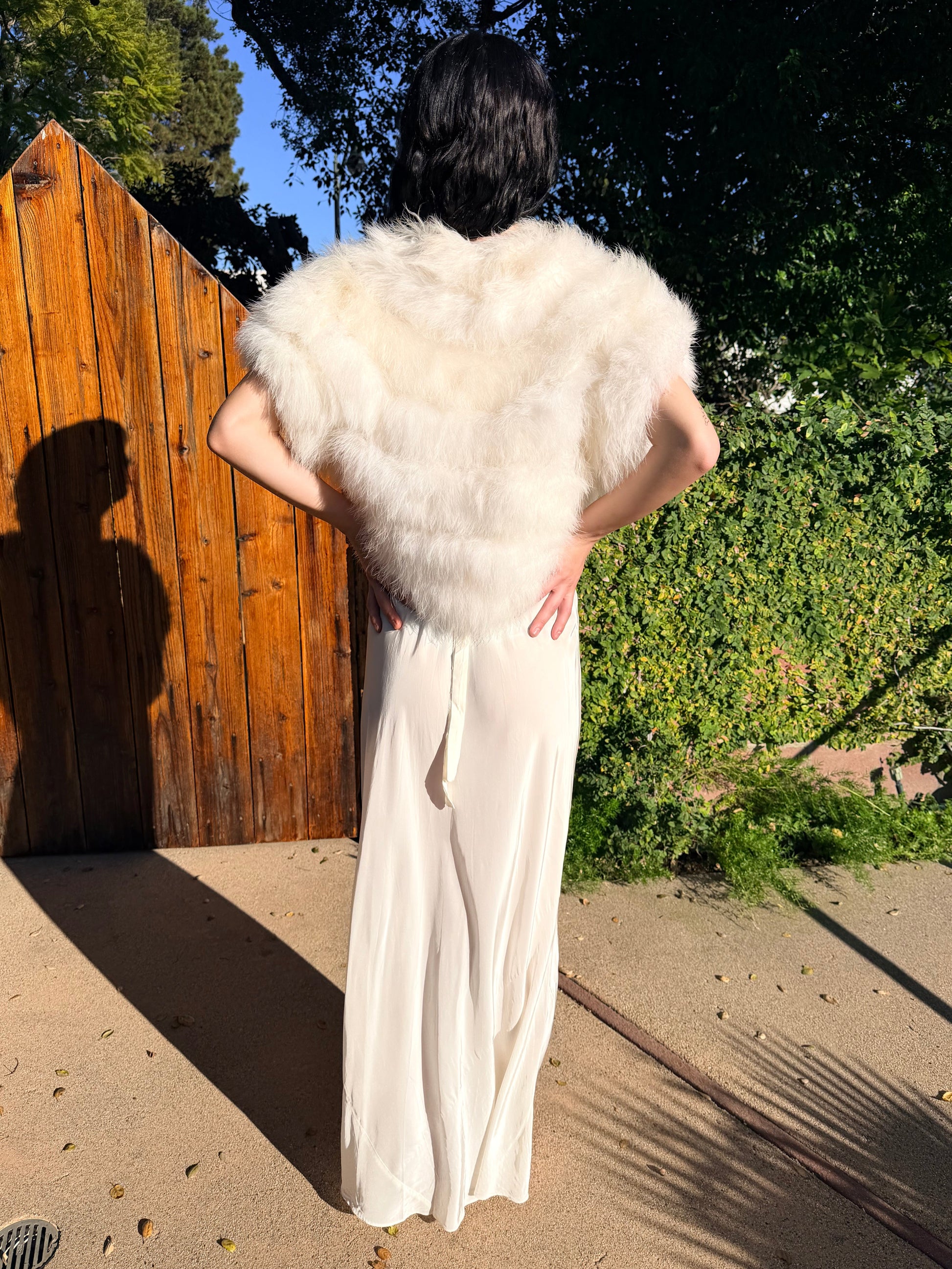 Person wearing a white fur coat and long white dress standing on a wooden platform with trees in the background.