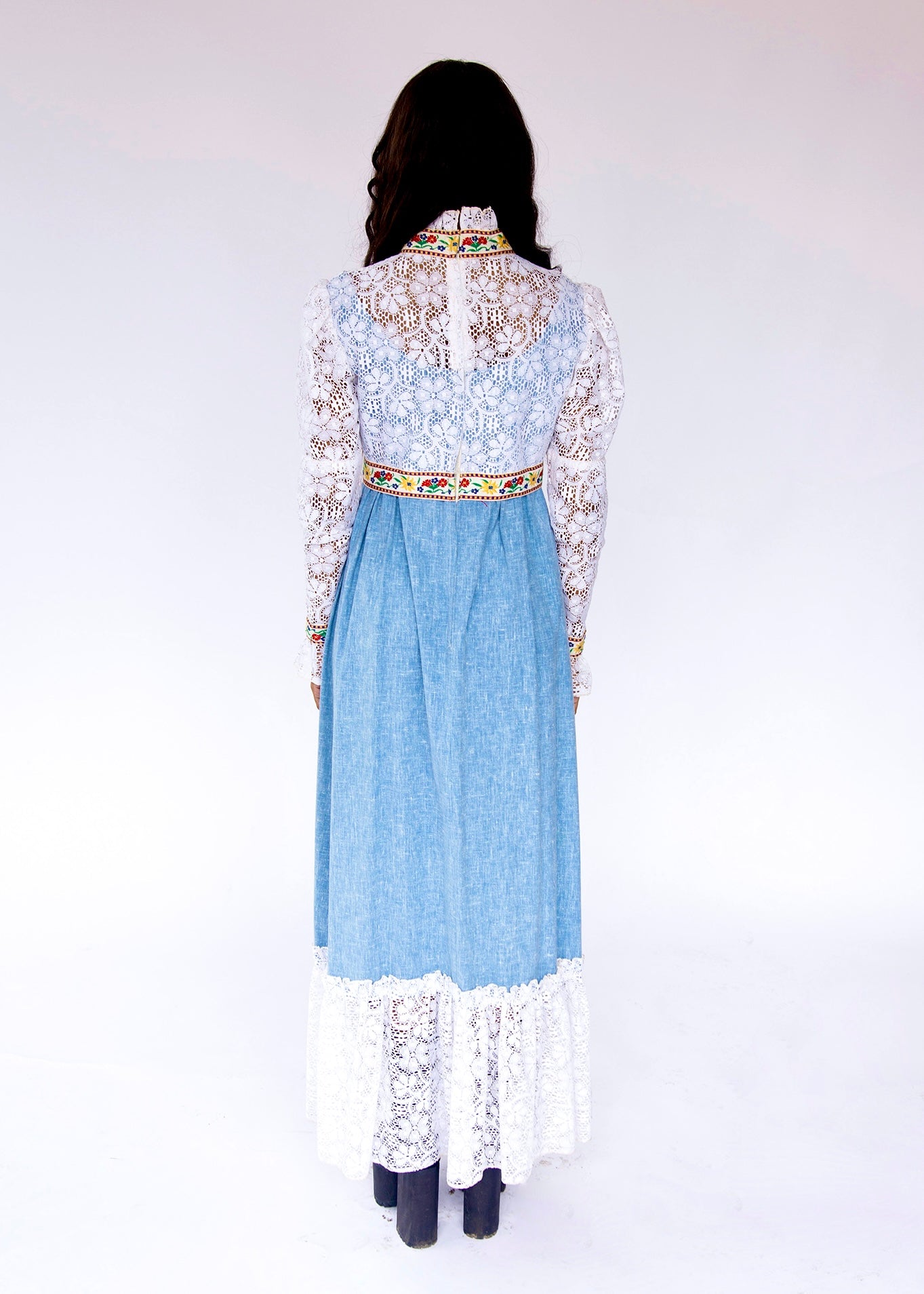 style boho hippie maxi dress made from denim and lace with mutton style long sleeve. Light blue denim maxi dress with intricate white lace overlays on the bodice and sleeves, a colorful patterned waistband, and a white lace hem, photographed against a plain white studio background.