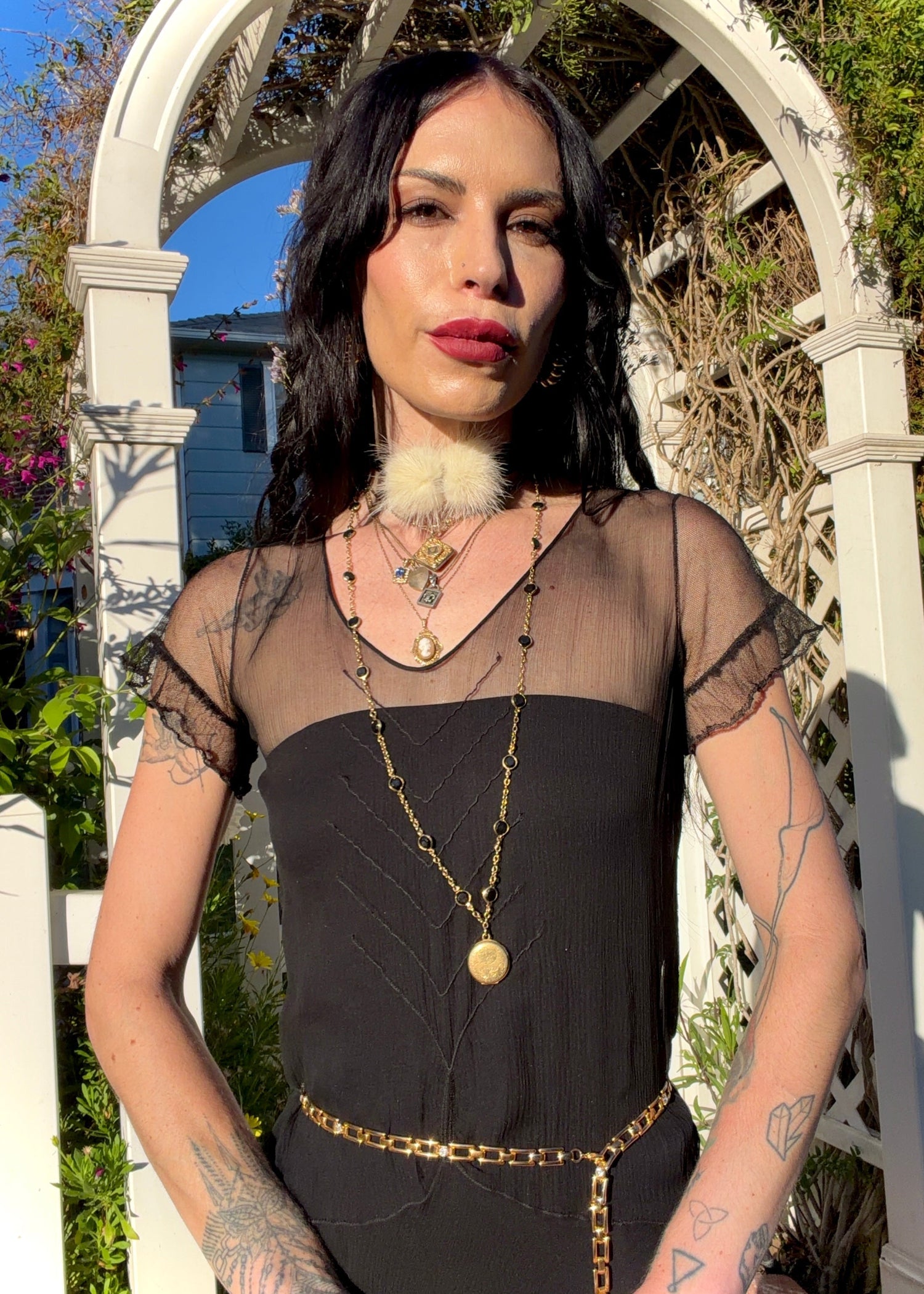 A woman stands outdoors under a white arched garden gate wearing a sheer black antique 1920's dress with a long black mesh skirt, layered gold necklaces, a gold chain belt, and beige high heels.