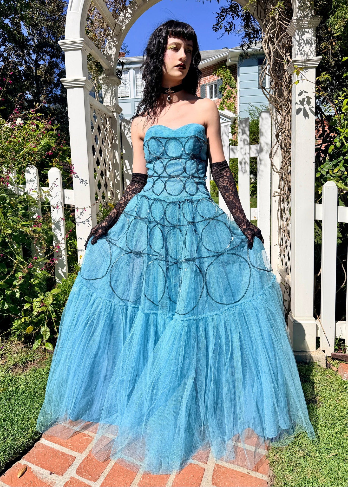 Woman in a 1950's blue strapless ball gown with black gloves standing in a garden.