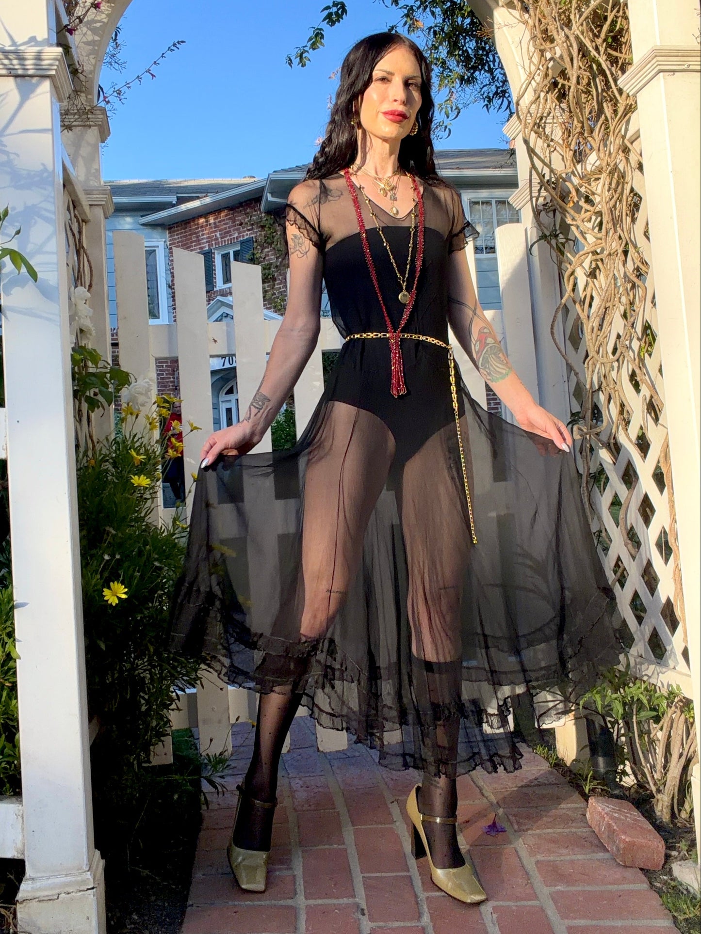 A woman stands outdoors under a white arched garden gate wearing a sheer black antique 1920's dress with a long black mesh skirt, layered gold necklaces, a gold chain belt, and beige high heels.