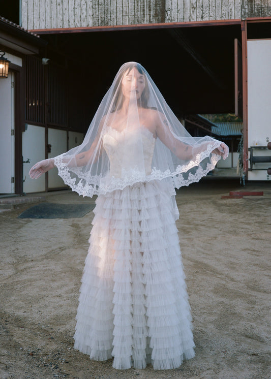 Woman in a vintage wedding dress with a veil standing in front of a rustic building.