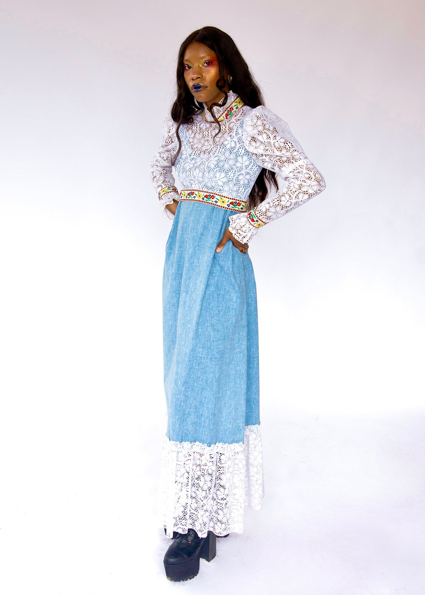 style boho hippie maxi dress made from denim and lace with mutton style long sleeve. Light blue denim maxi dress with intricate white lace overlays on the bodice and sleeves, a colorful patterned waistband, and a white lace hem, photographed against a plain white studio background.