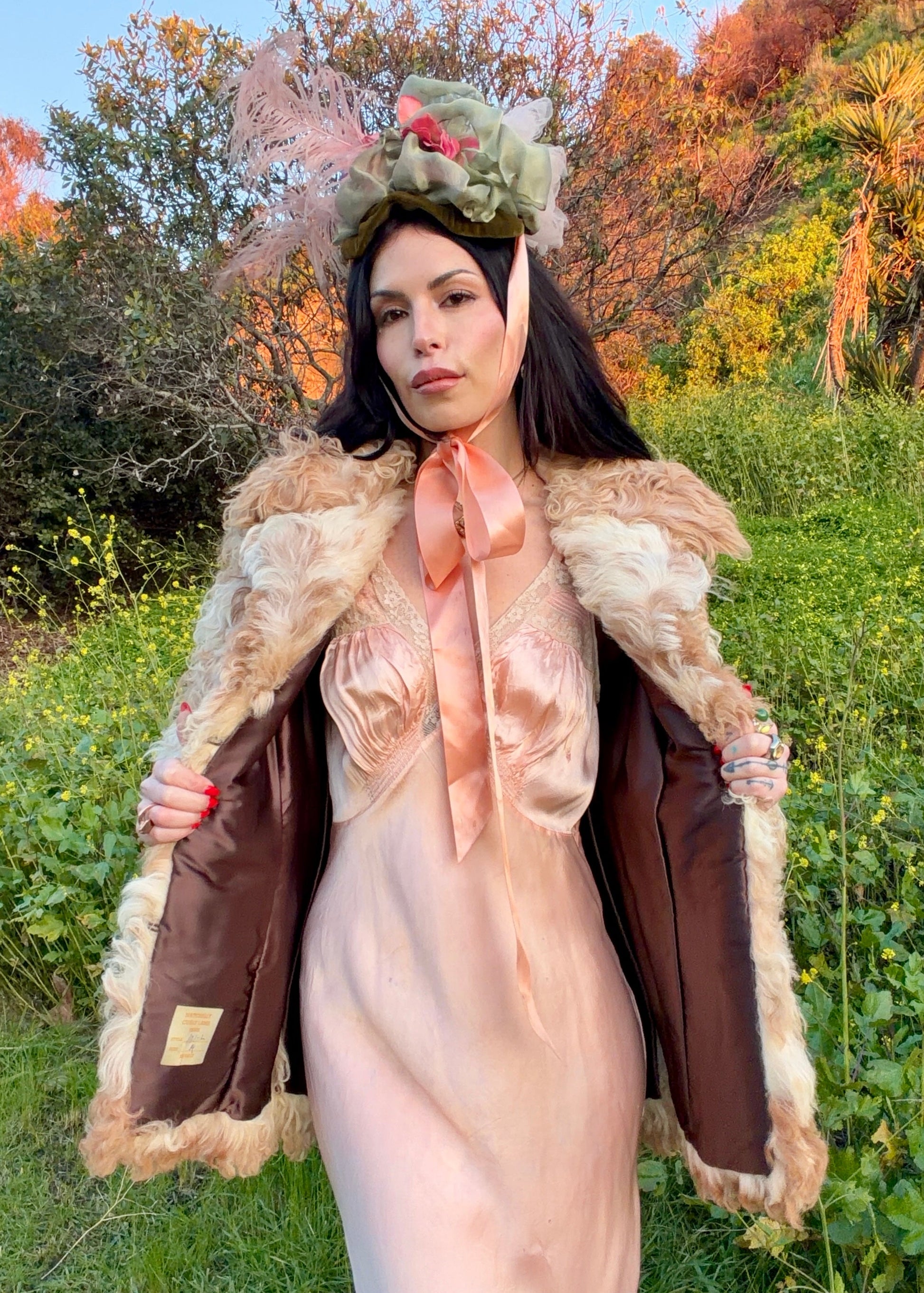 woman wearing a 1970's curly Mongolian and Tibetan sheep's fur coat in fluffy beige and brown colors in a natural setting with trees and grass.