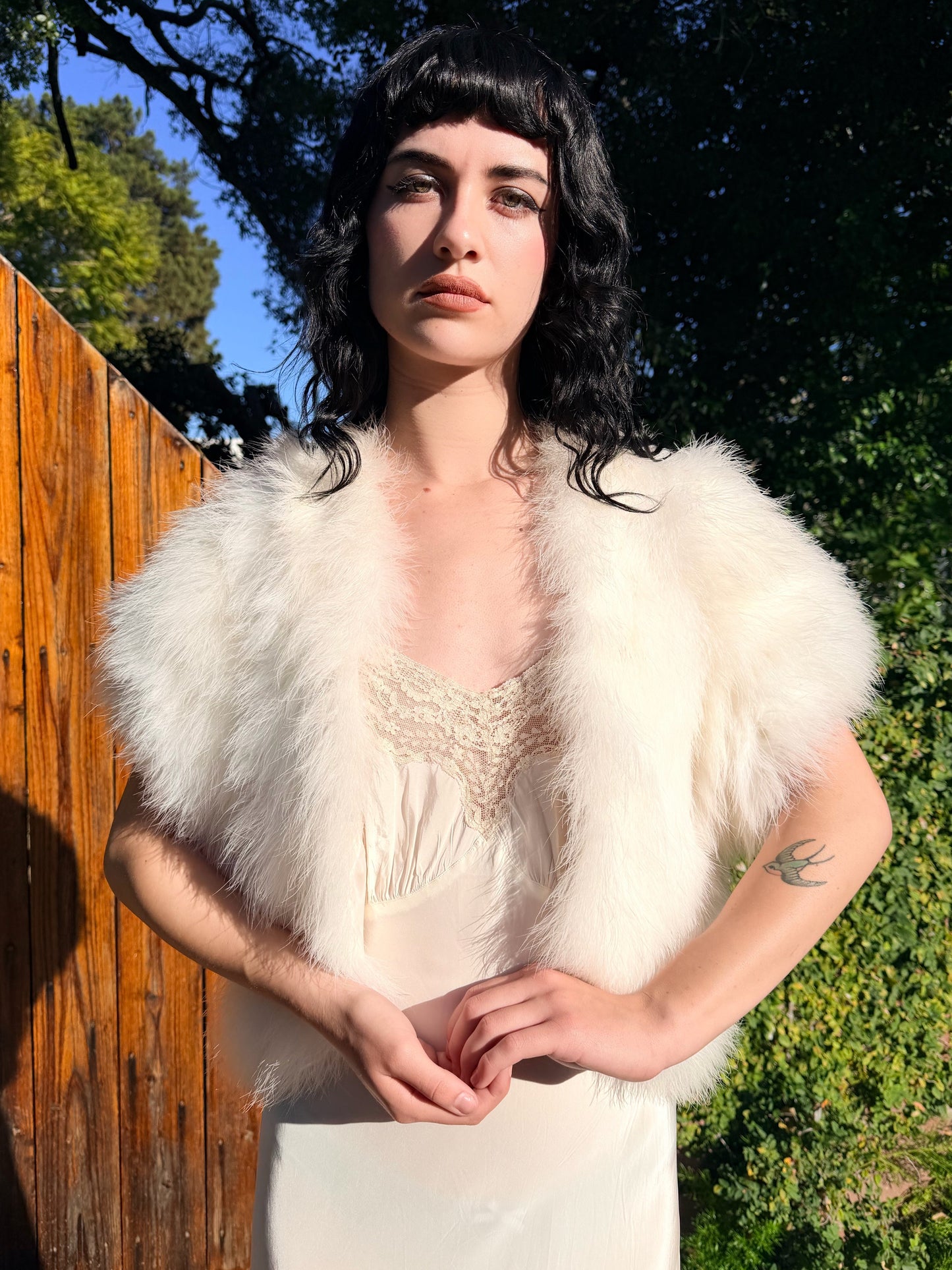 Person wearing a white marabou vest outdoors with trees and a wooden fence in the background