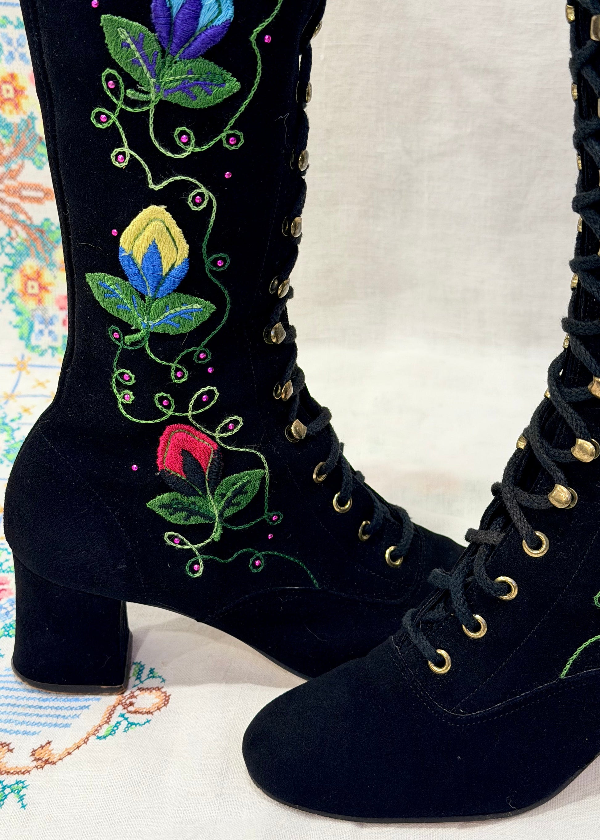 Black knee-high 1960's suede penny lane gogo boots with colorful floral embroidery on a decorative background