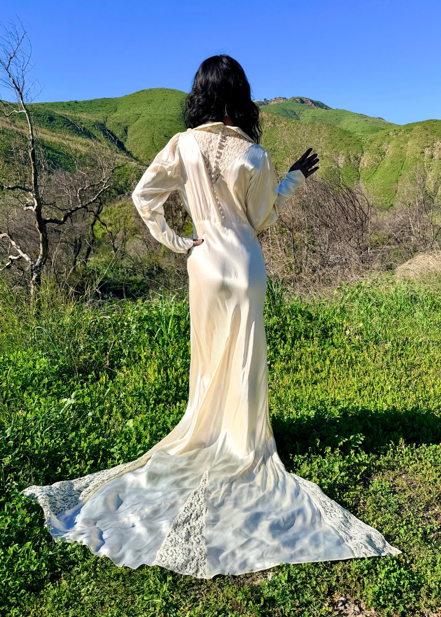 Person in a long, light-colored dress standing in a natural setting with green hills and blue sky.